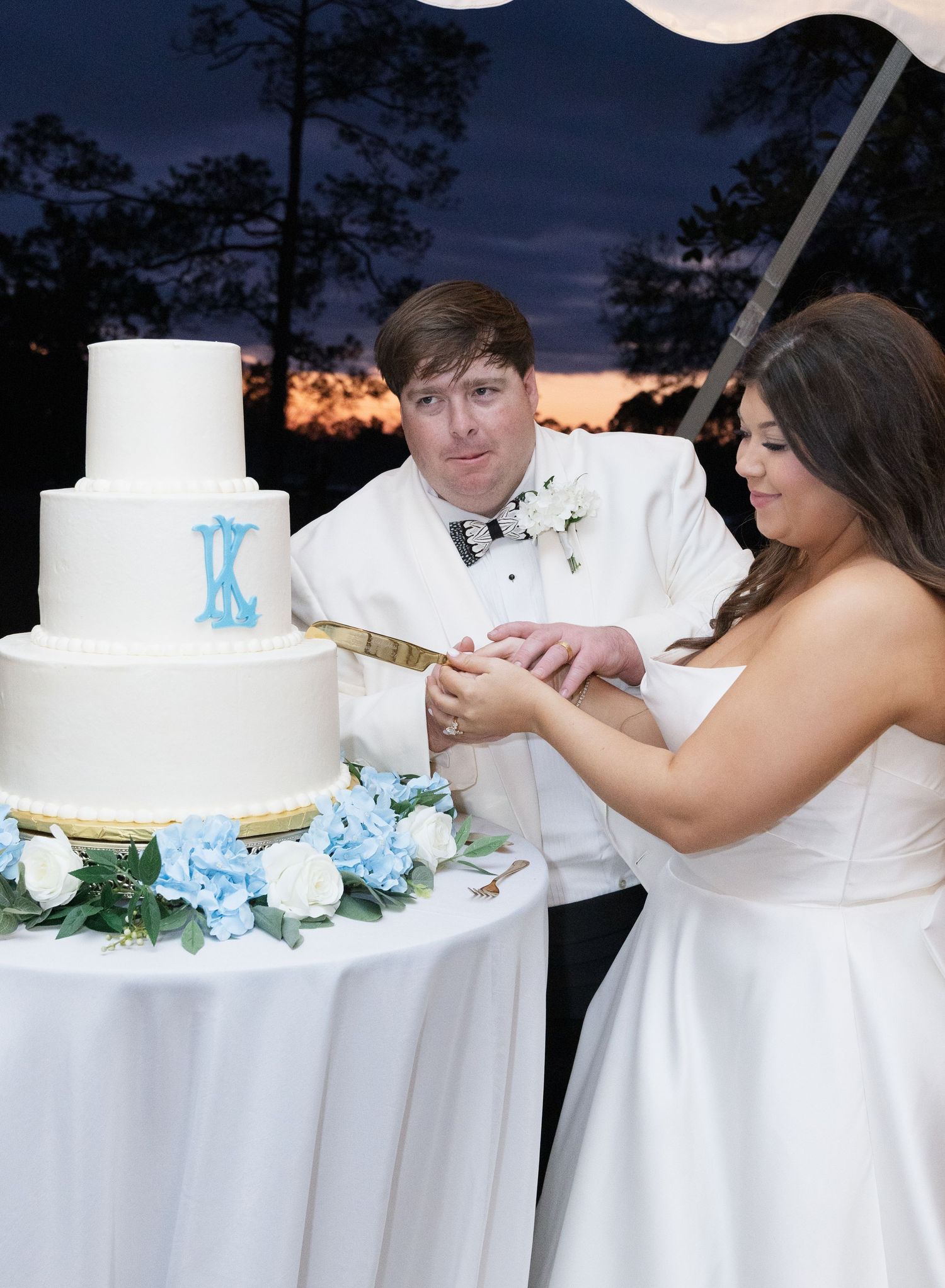 Bride and groom cut white three-tier wedding cake with blue monogram at outdoor evening reception.