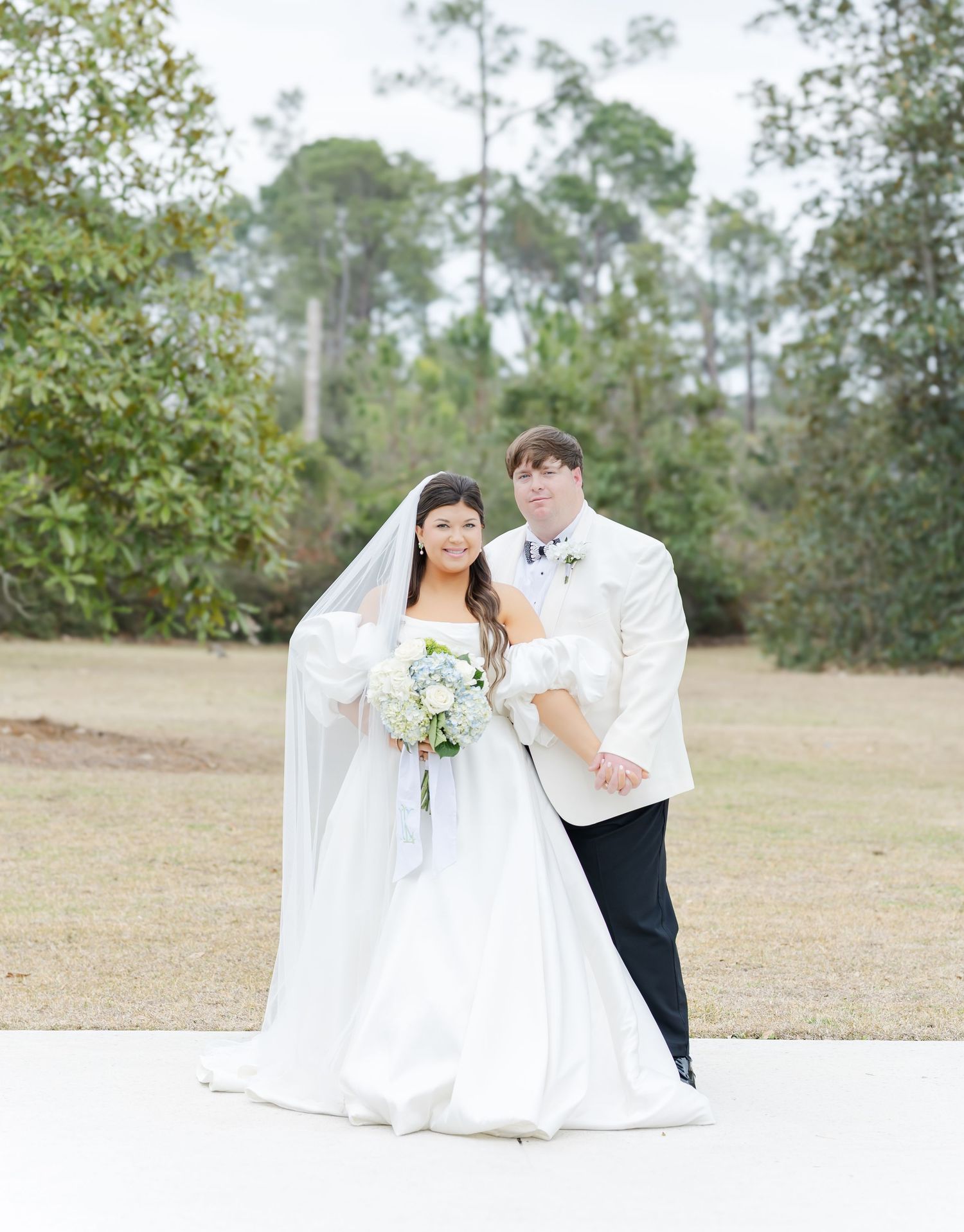Bride and groom pose together outdoors, bride in strapless gown with veil holding white floral bouquet.