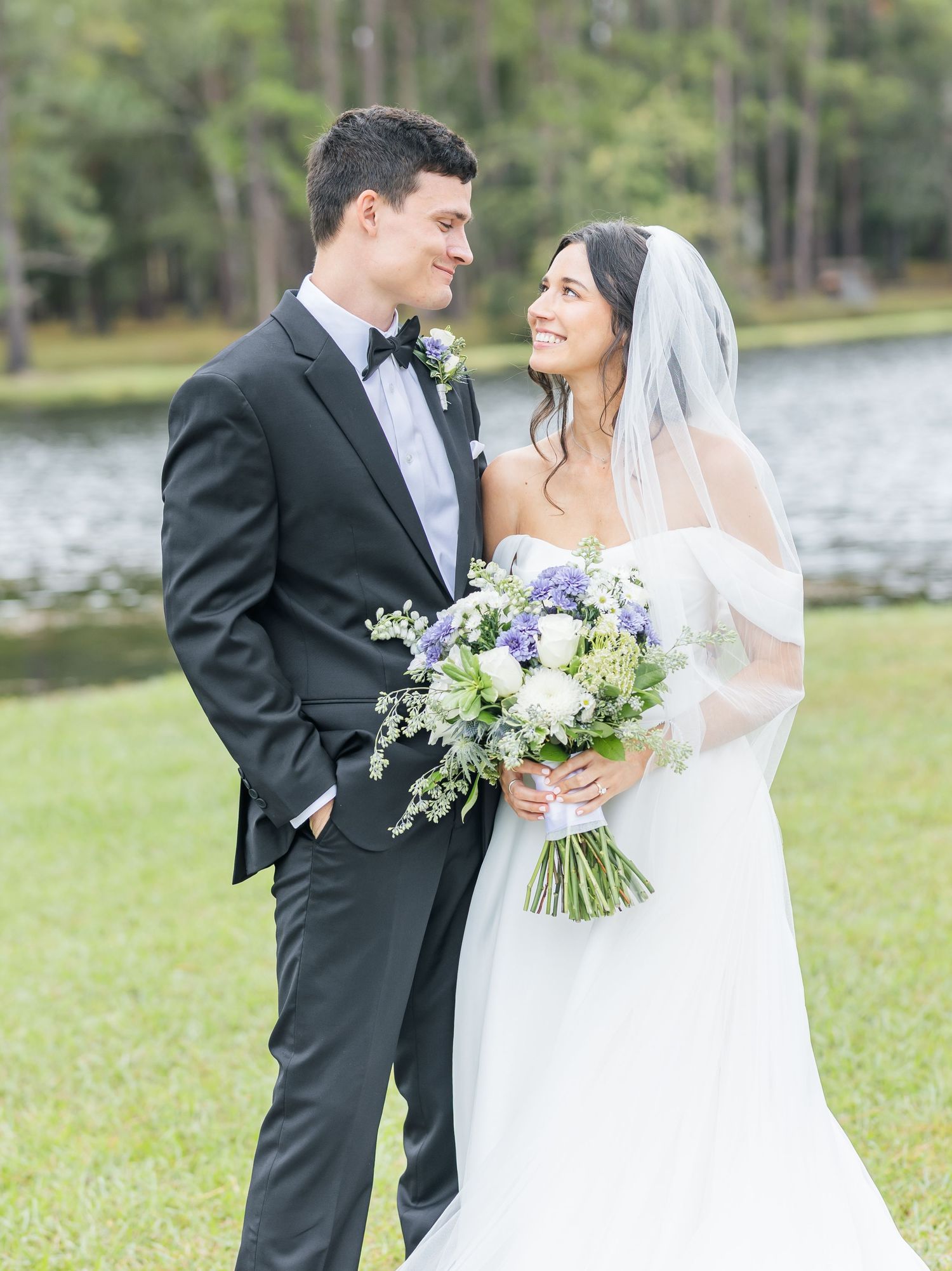 A romantic wedding couple poses outdoors near a lake with elegant floral bouquets in classic black and white attire.