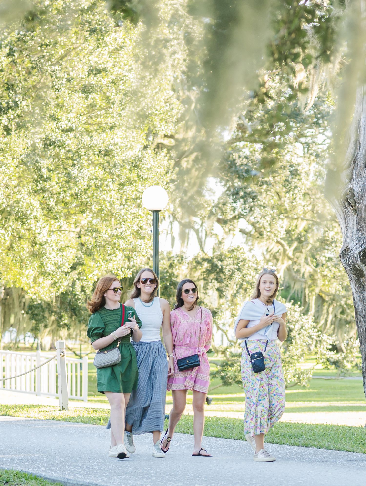 Four friends walking together on a sunny path through a lush park with trees and lamp posts.