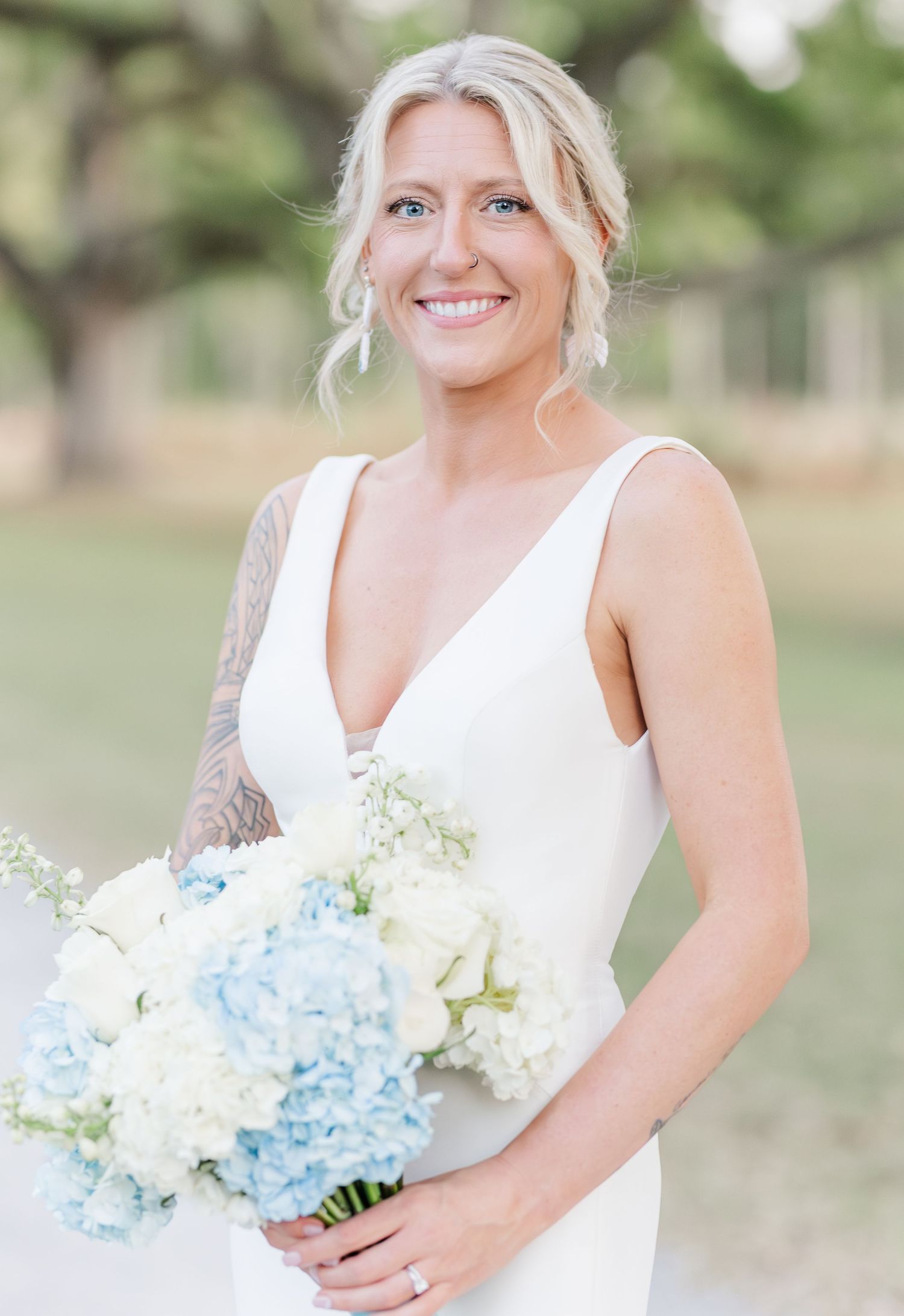 Smiling blonde bride in sleeveless white gown holding blue and white hydrangea bouquet outdoors.