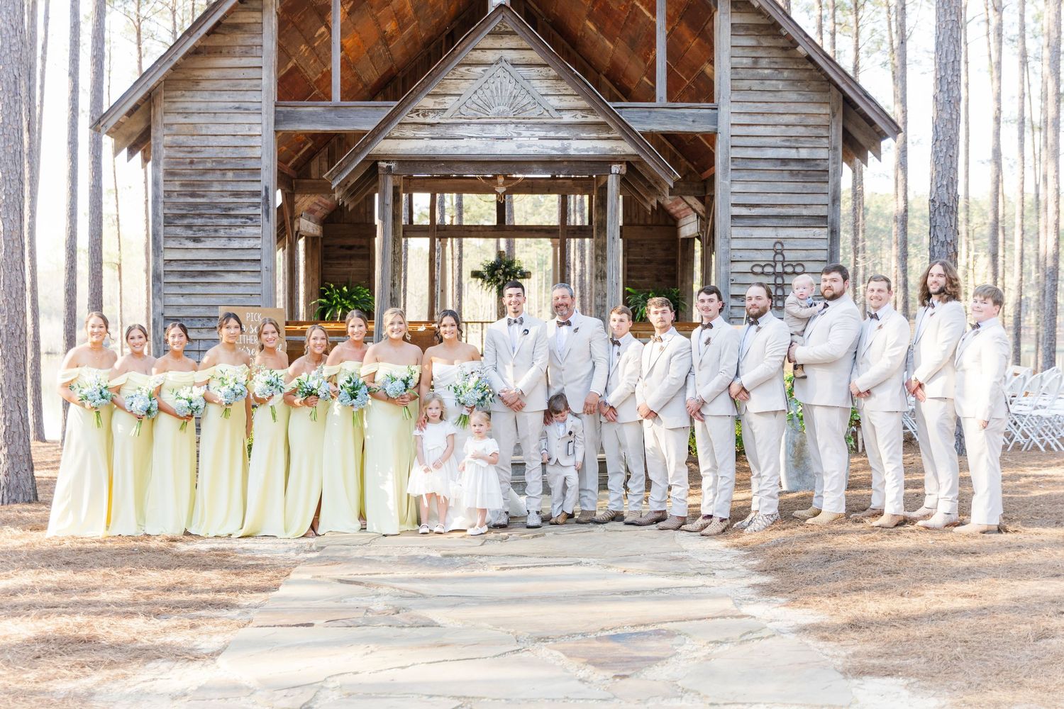 Wedding party posed before rustic wooden chapel with bridesmaids in yellow and groomsmen in tan suits.
