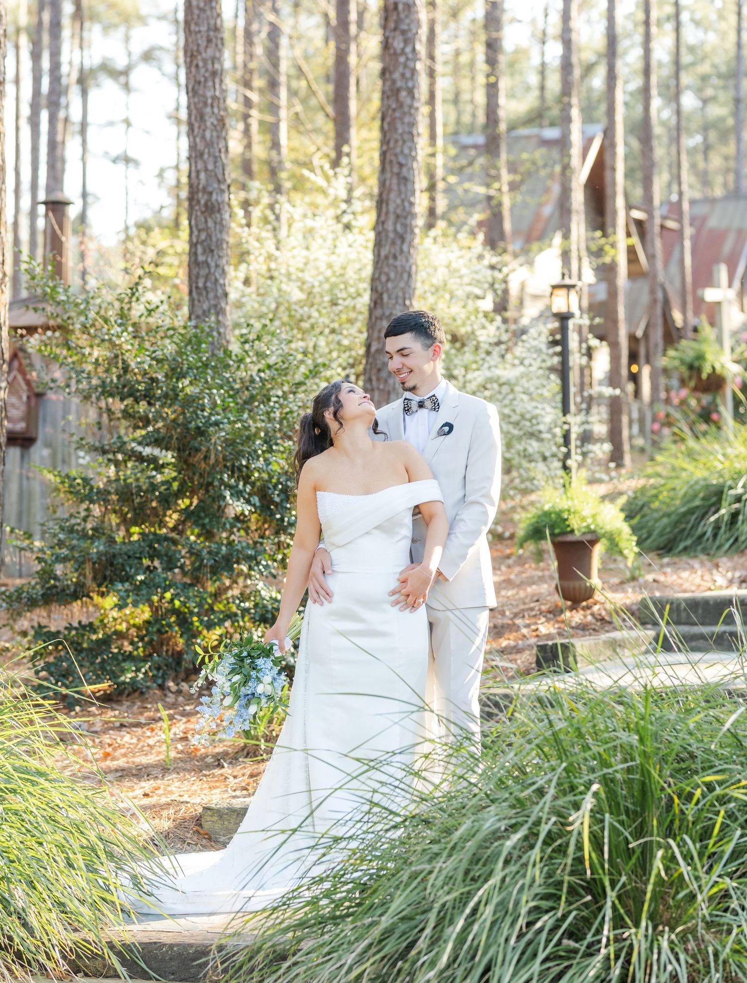 Bride in strapless white gown and groom in tan suit share a romantic moment in a lush garden wedding setting.