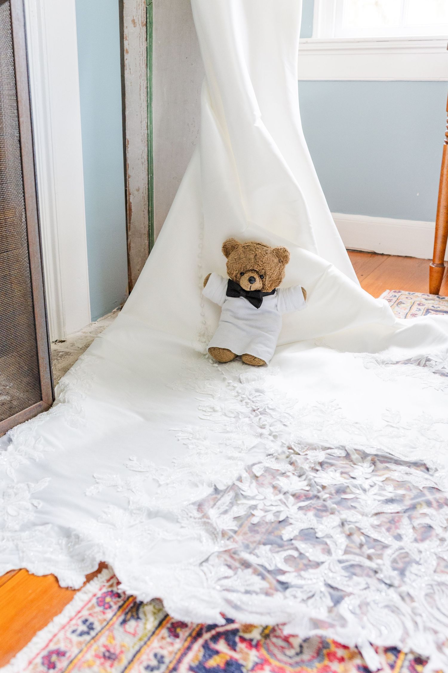 A brown teddy bear in a tuxedo shirt sits beneath a flowing white wedding dress train on a hardwood floor.