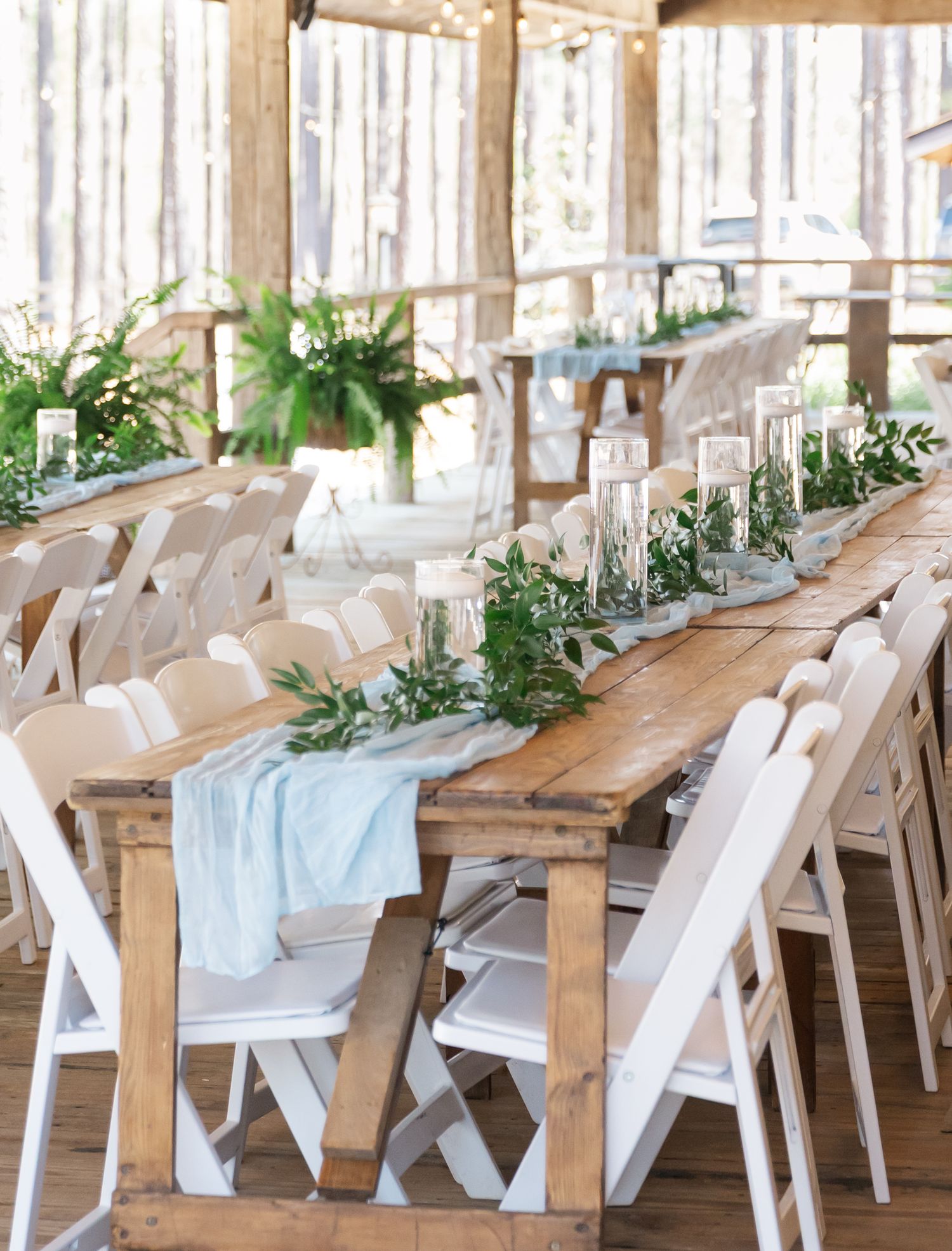 Rustic wooden farm tables with white chairs, greenery runners, and candles in an open-air barn venue.