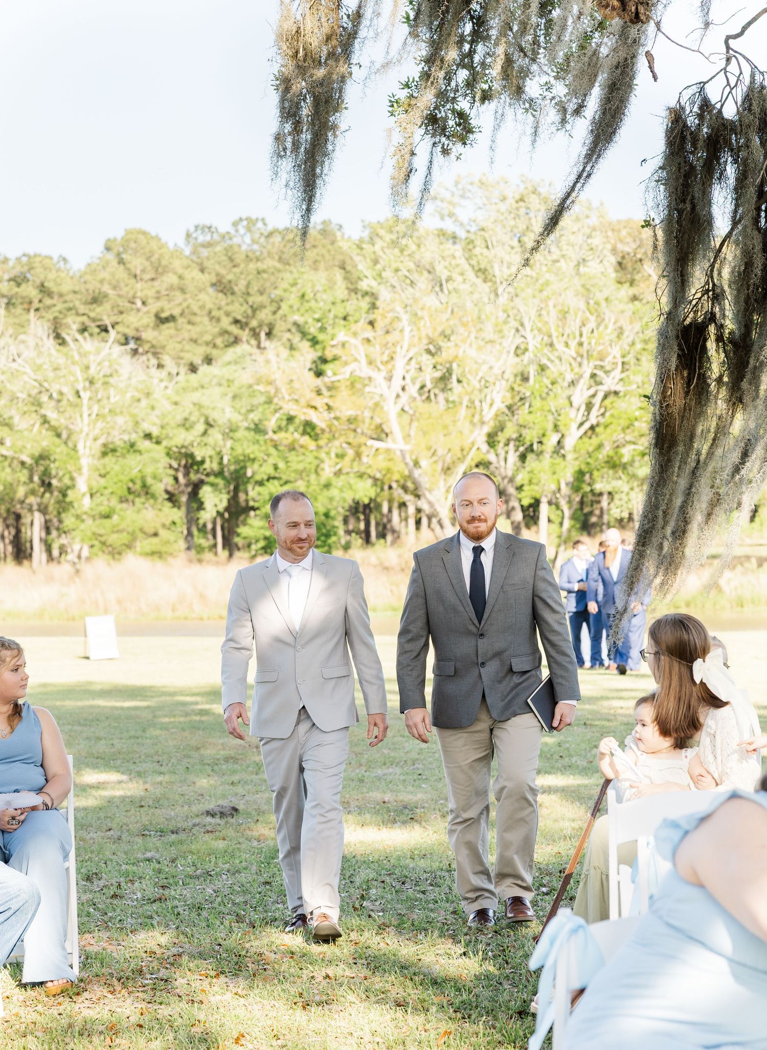 Groom in gray suit walks down outdoor aisle with groomsman at sunny wedding ceremony.