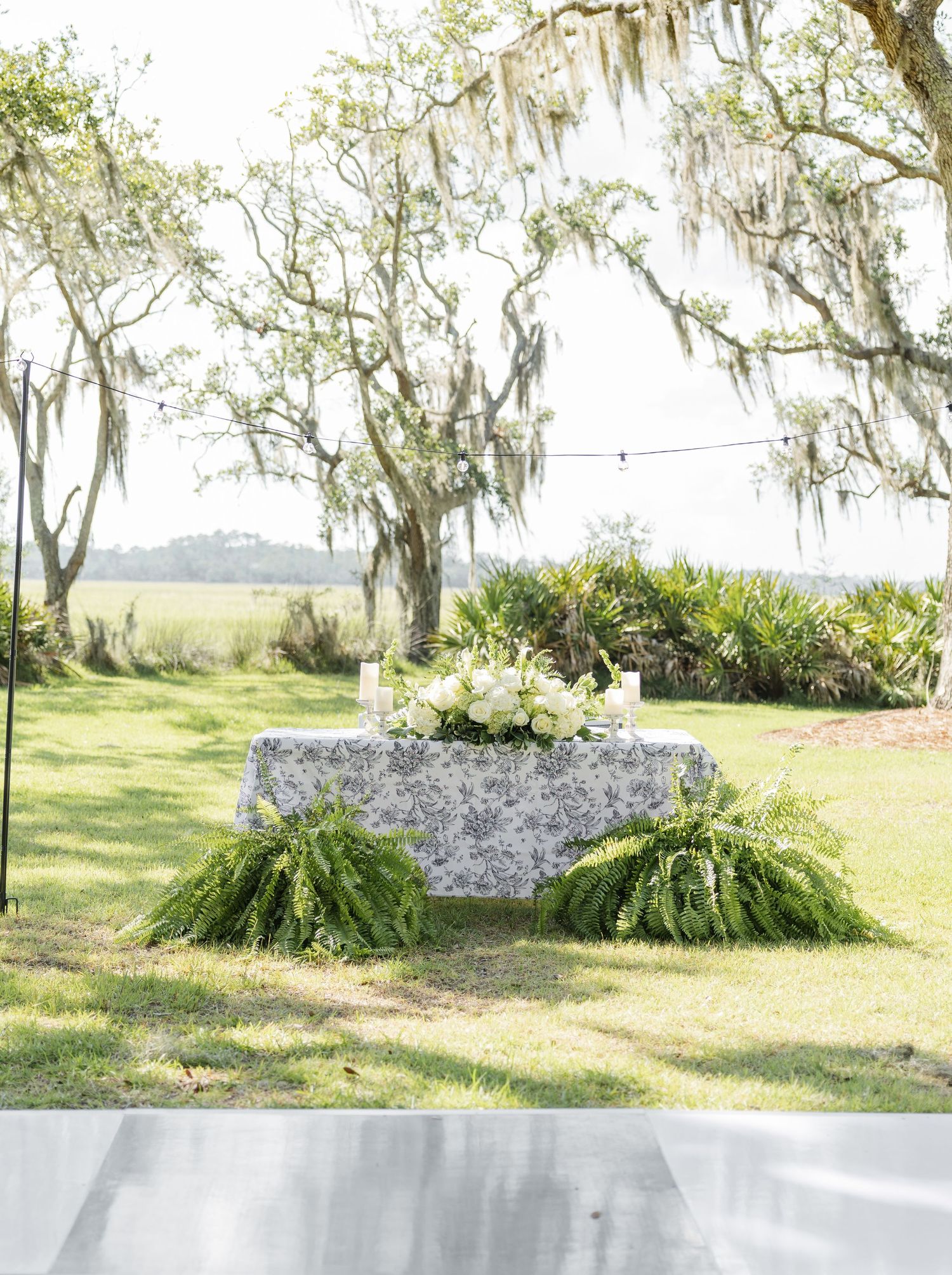 Outdoor wedding sweetheart table with blue toile linen, white floral centerpiece, and ferns under Spanish moss-draped oak trees.