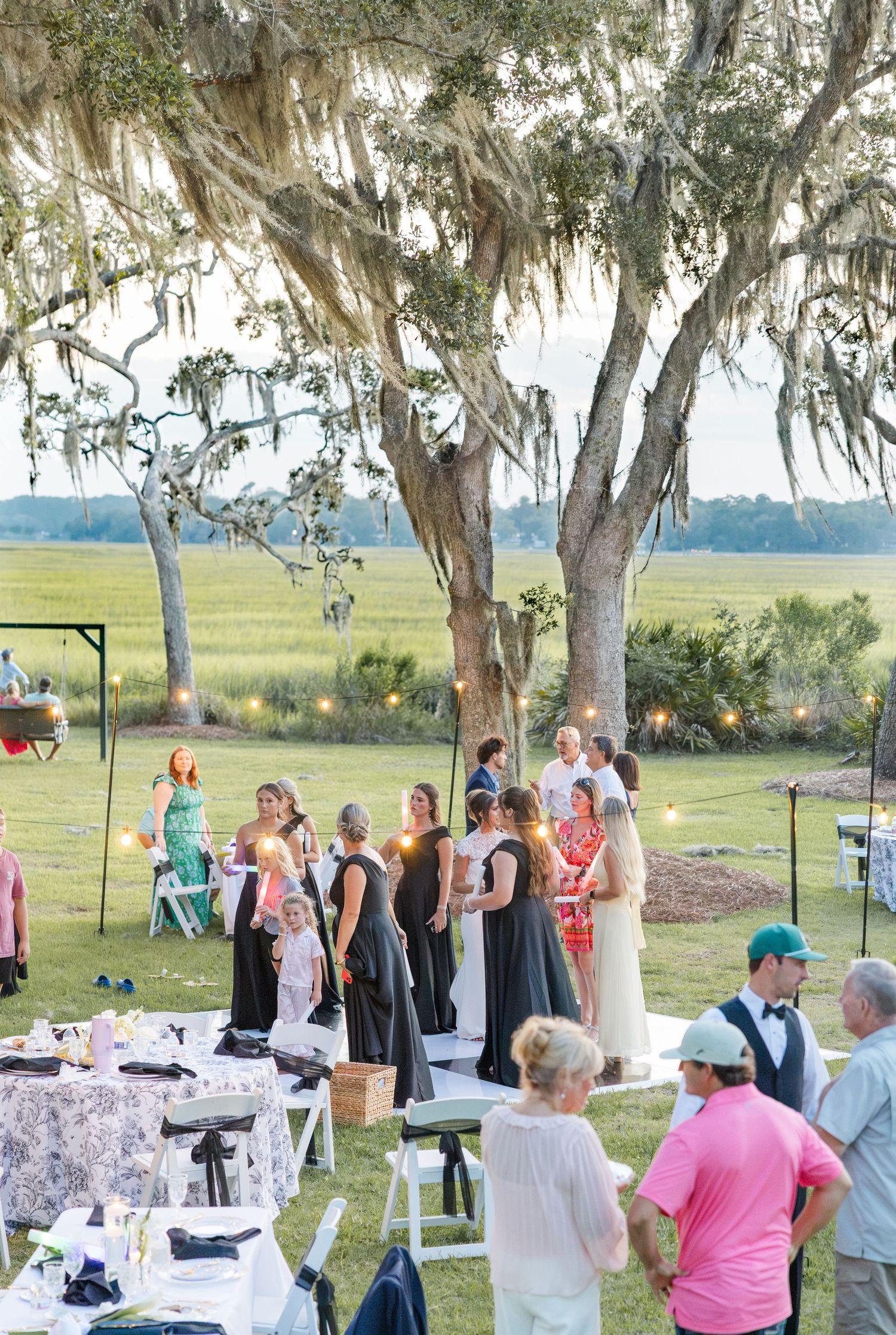 Wedding guests mingle under Spanish moss-draped oak trees at an elegant outdoor waterfront reception at golden hour.