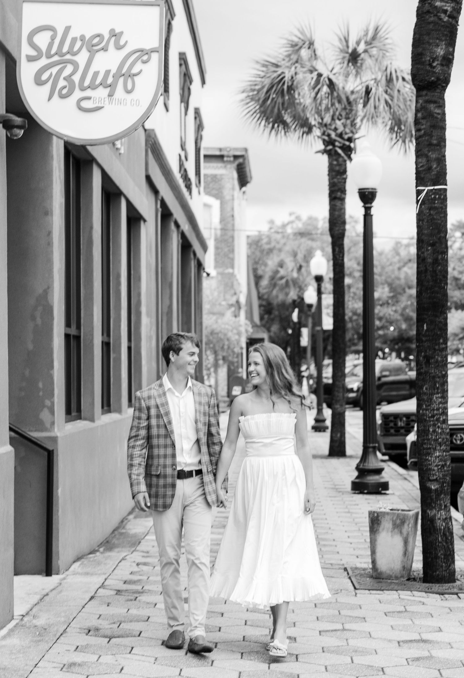 Couple holds hands walking past Silver Bluff cafe on brick sidewalk with palm trees in black and white photo.
