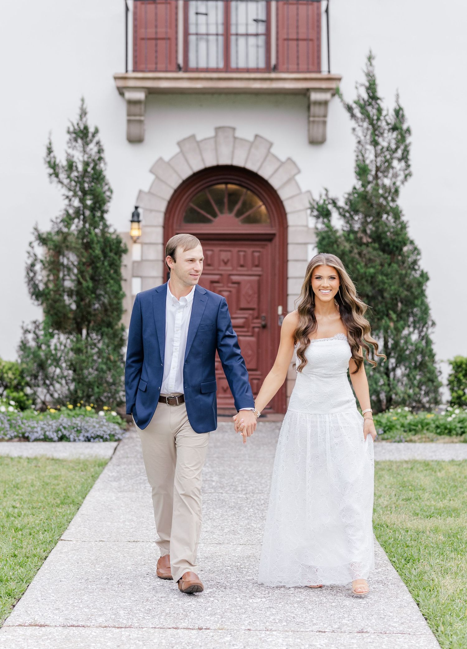 Couple holding hands walking down a pathway in front of a white building with a red arched door.