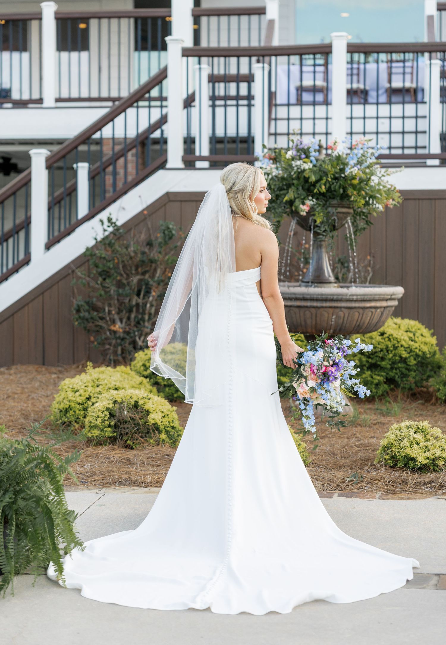 Bride in strapless white gown with long veil holds colorful bouquet with back to camera at venue.