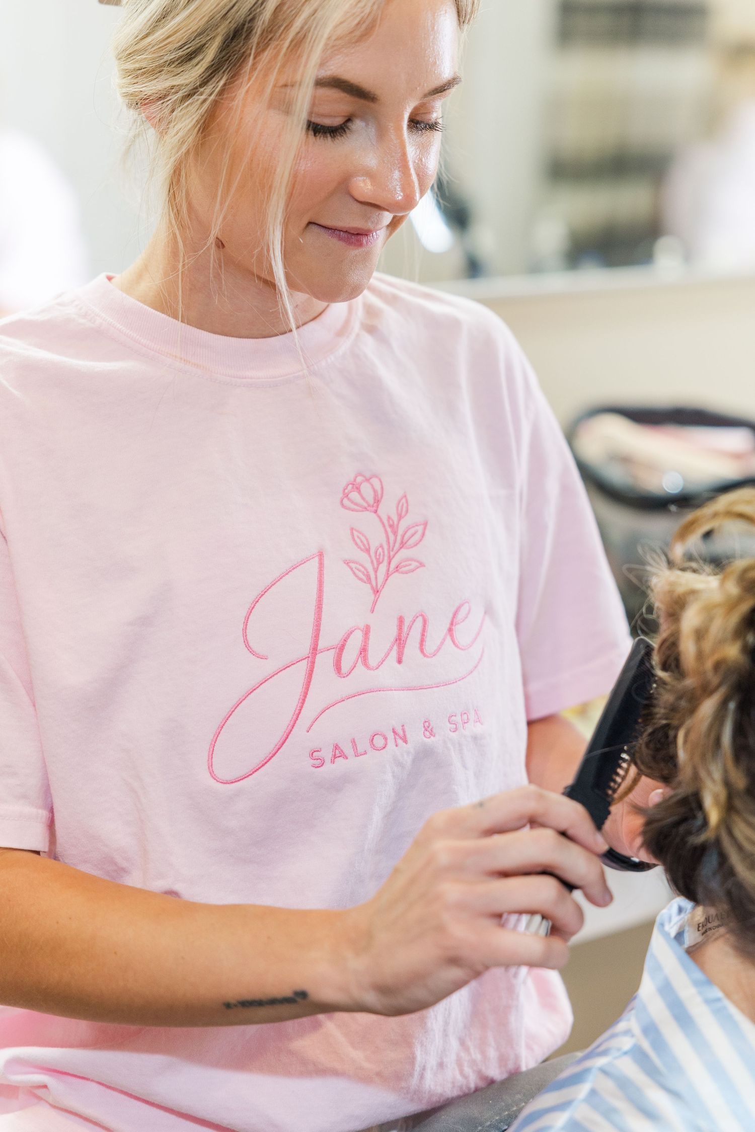 A smiling hairstylist in a pink Jane Salon & Spa shirt styling a client's hair in a bright salon.
