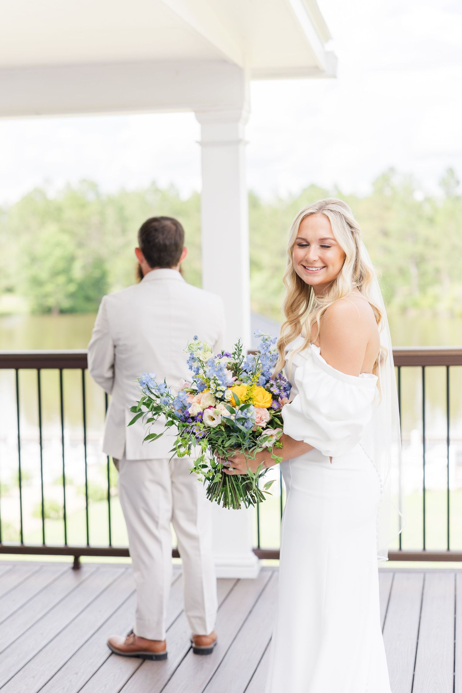 Smiling bride holding colorful blue and yellow bouquet before first look with groom on lakeside wedding deck.
