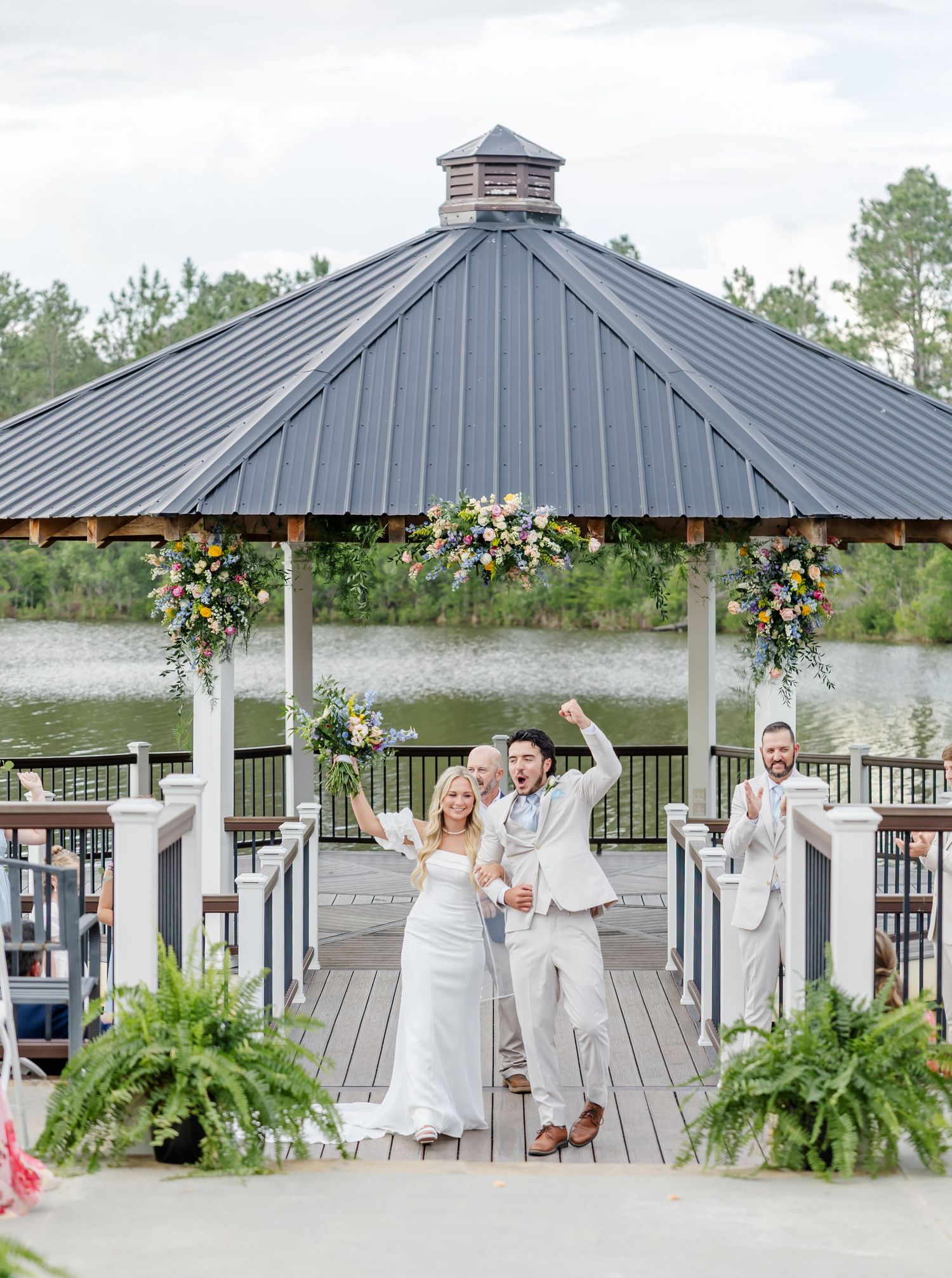 Jubilant newlyweds celebrate with raised arms at gazebo ceremony venue overlooking scenic lake.