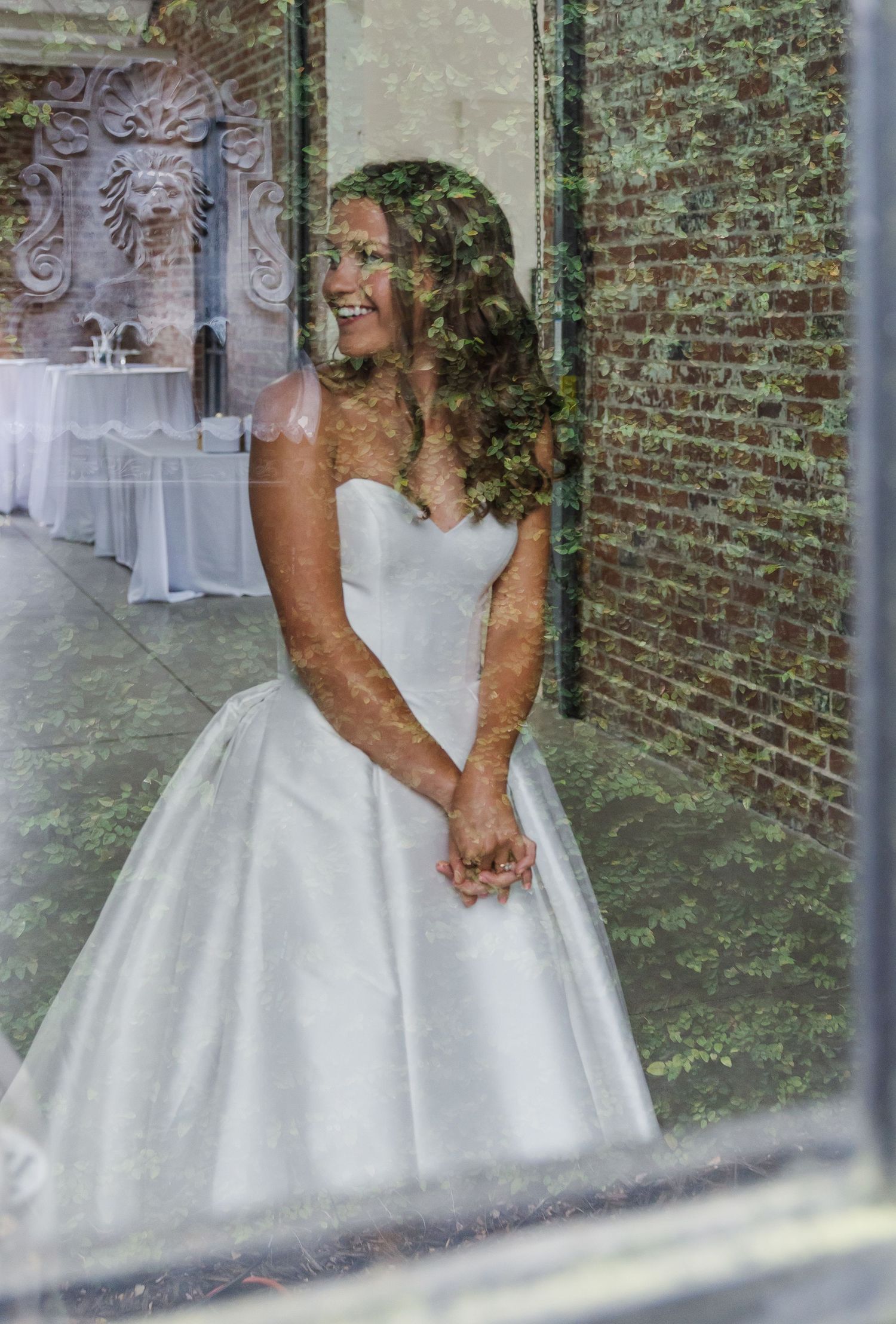 Bride in white ball gown smiling, photographed through a window with brick wall backdrop.