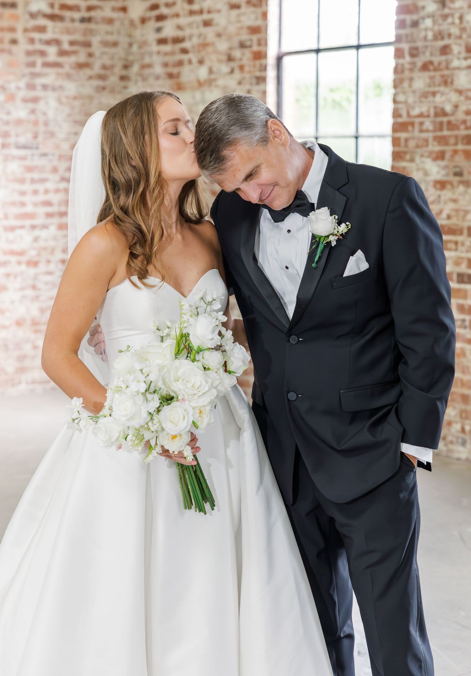 Bride kisses father on cheek while holding white bouquet at wedding venue.