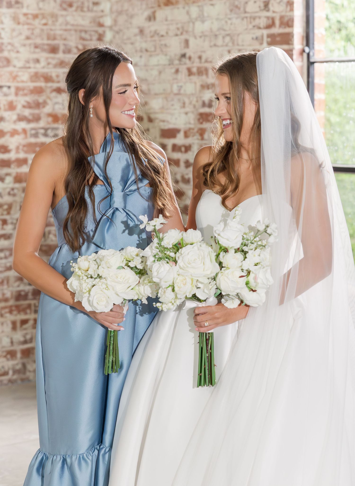 Bridesmaid in dusty blue halter gown and bride in white strapless gown holding white rose bouquets by brick wall.
