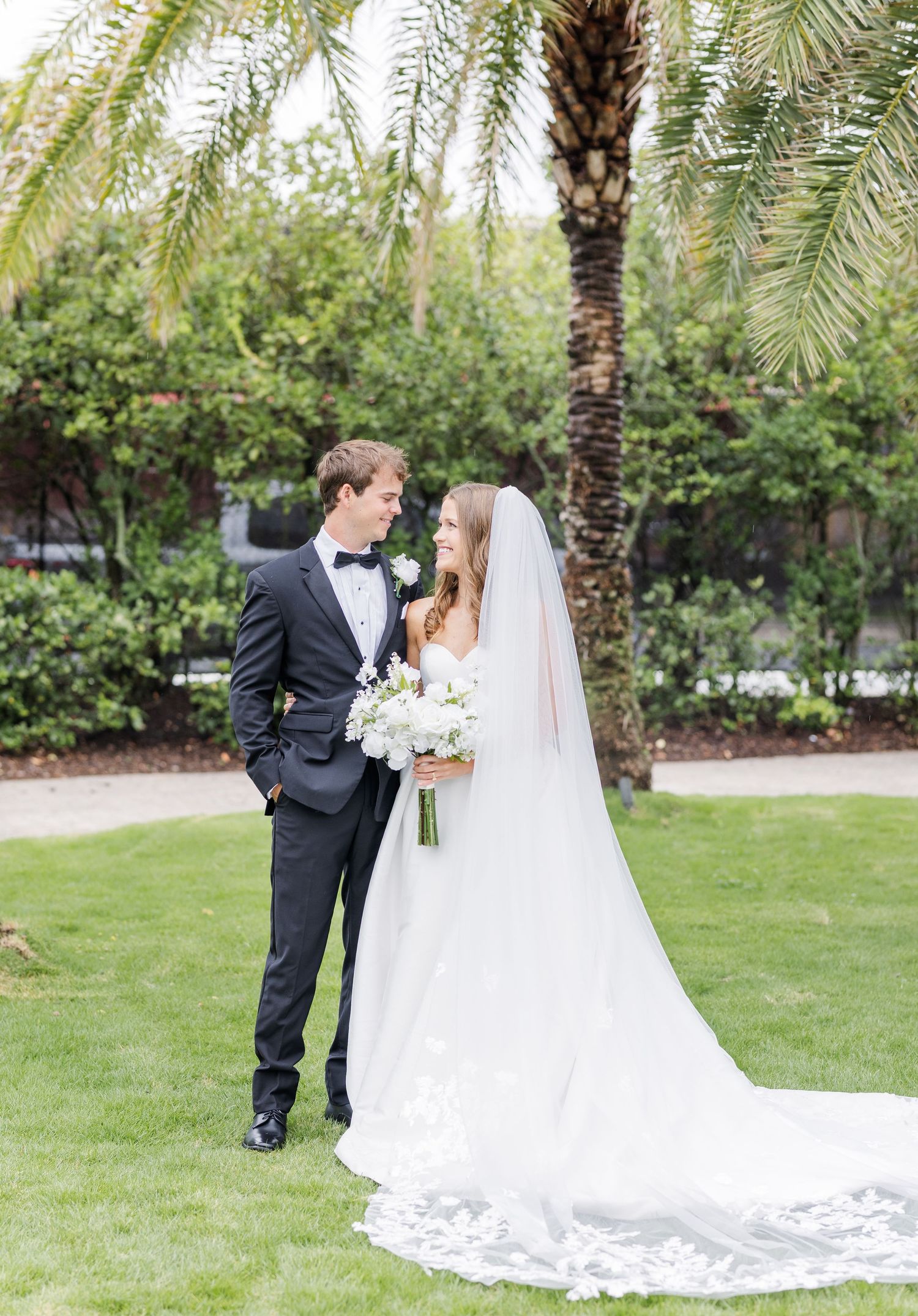 Bride and groom sharing a romantic gaze outdoors on green lawn with palm tree backdrop.
