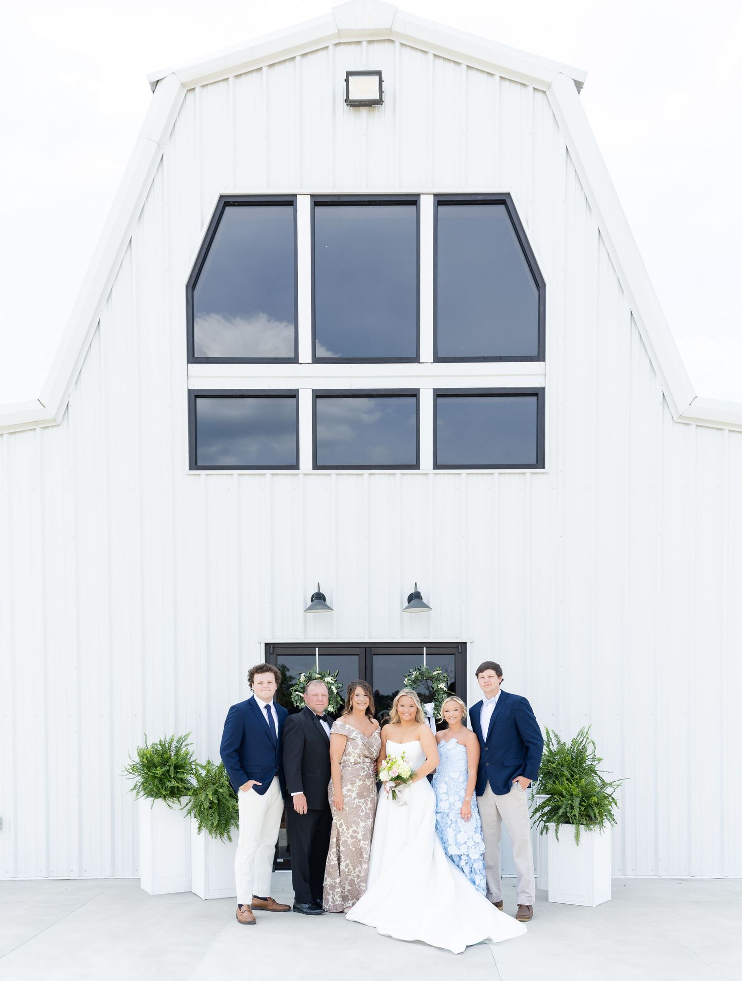 Wedding party poses in front of a white barn venue with large windows and lush greenery planters.