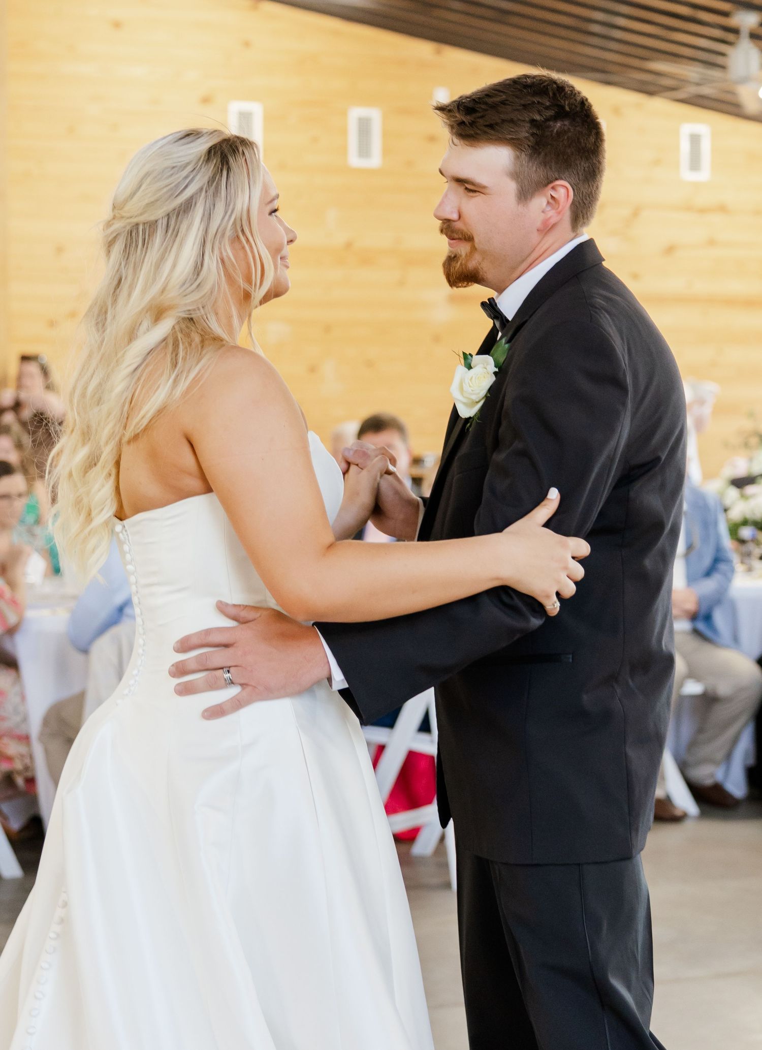 Bride and groom sharing first dance, smiling at each other in rustic wood-paneled reception venue.