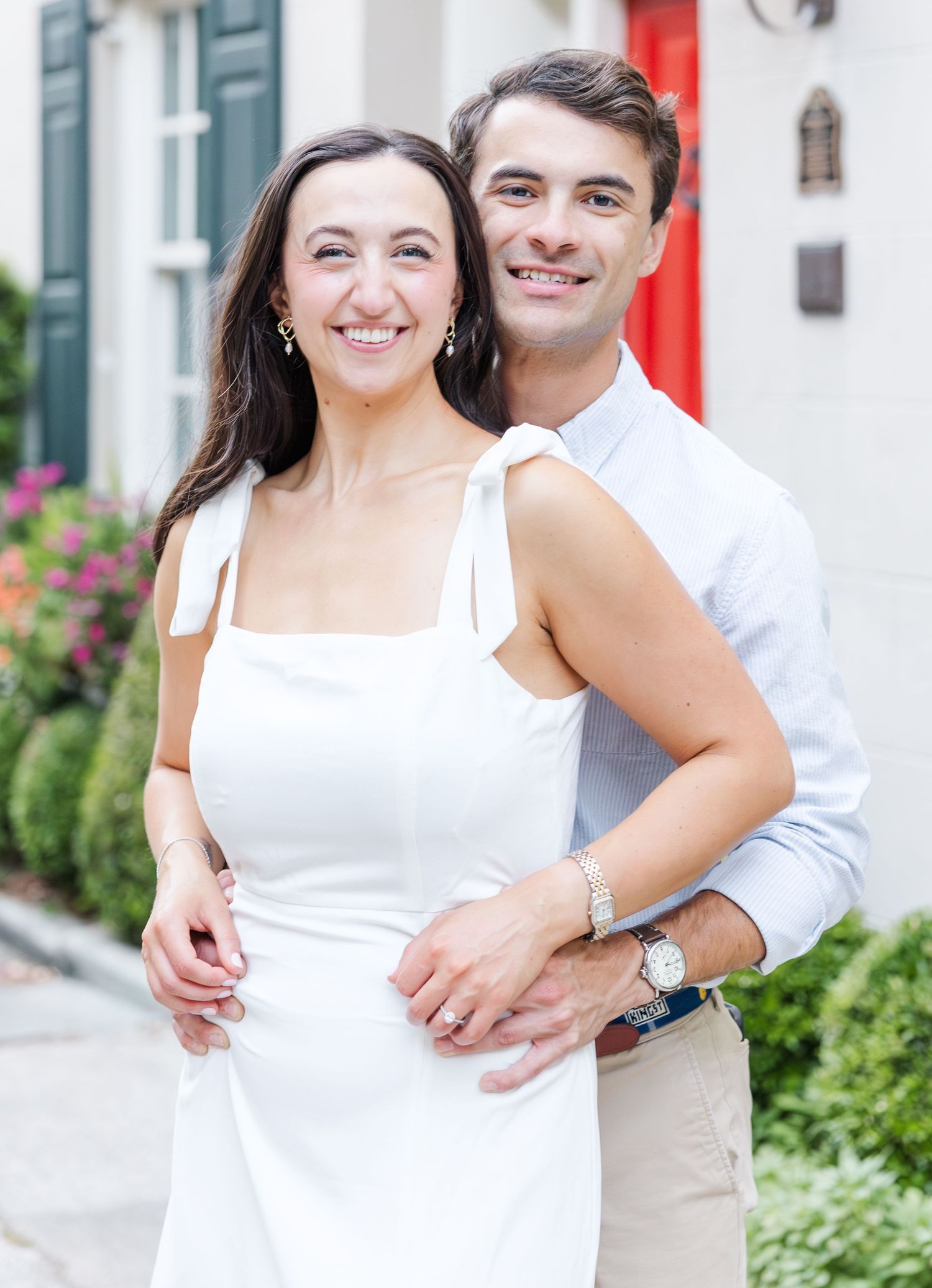 Smiling couple embracing closely outdoors in front of a red door with colorful flowers nearby.