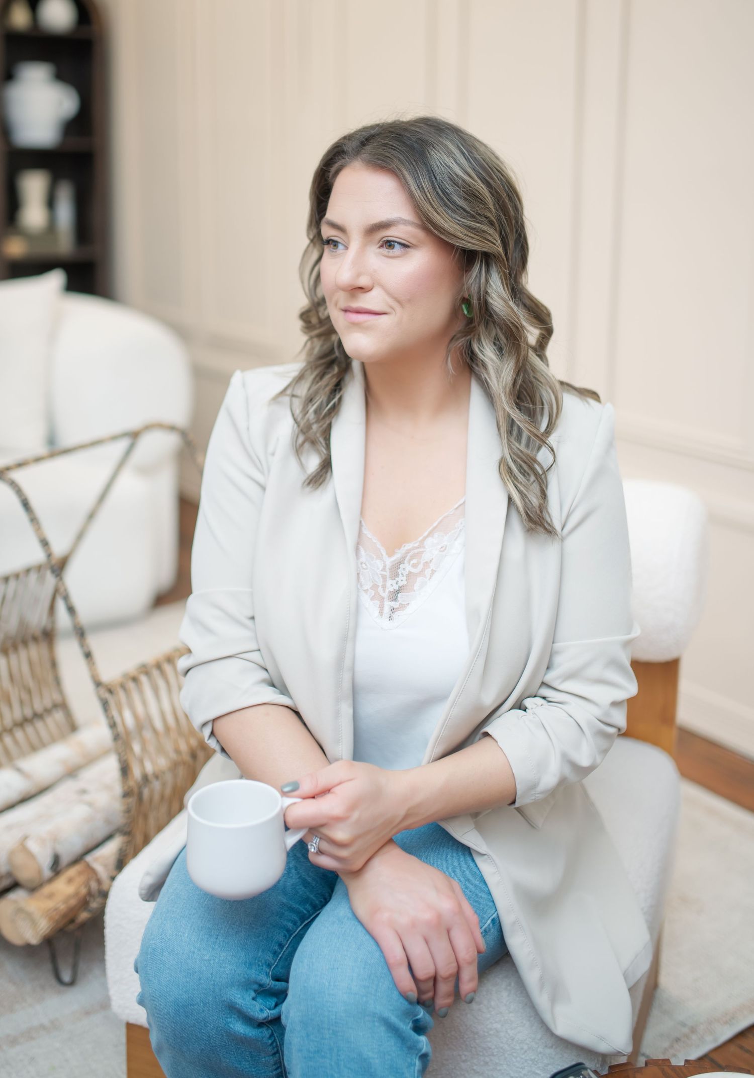 Professional woman in light blazer and jeans sits casually in modern interior setting with natural lighting.