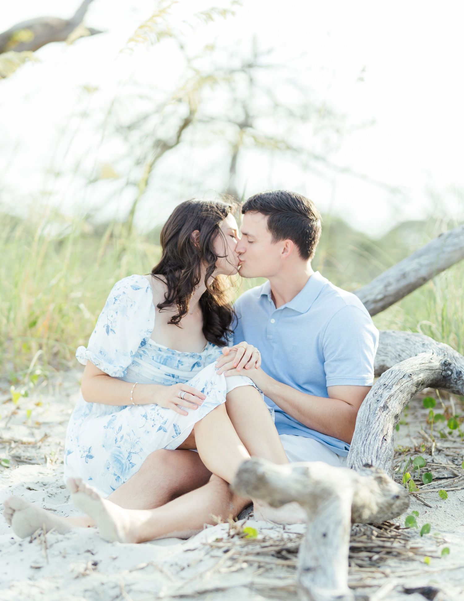 A couple in light blue and white sits together on a sandy beach near driftwood.