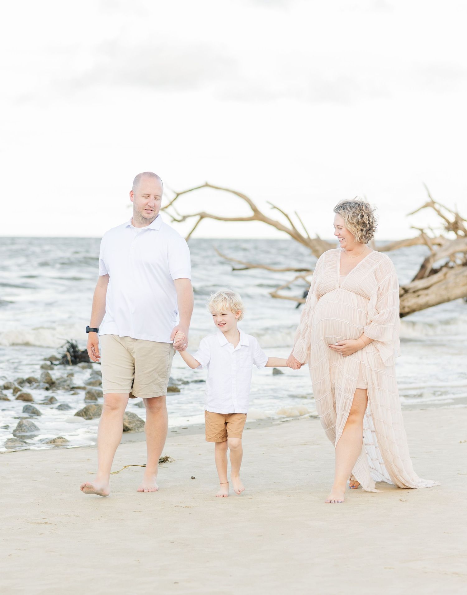 A family walks together along a scenic beach with driftwood and ocean waves in the background on a sunny day.
