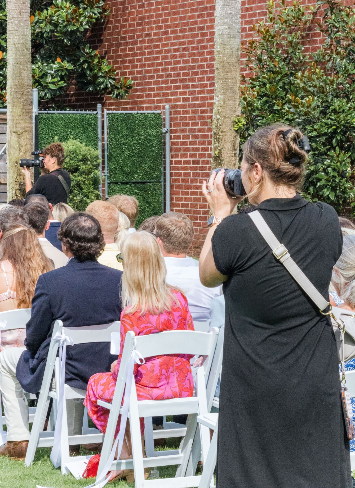 Several people seated in white folding chairs at an outdoor event next to a brick building.