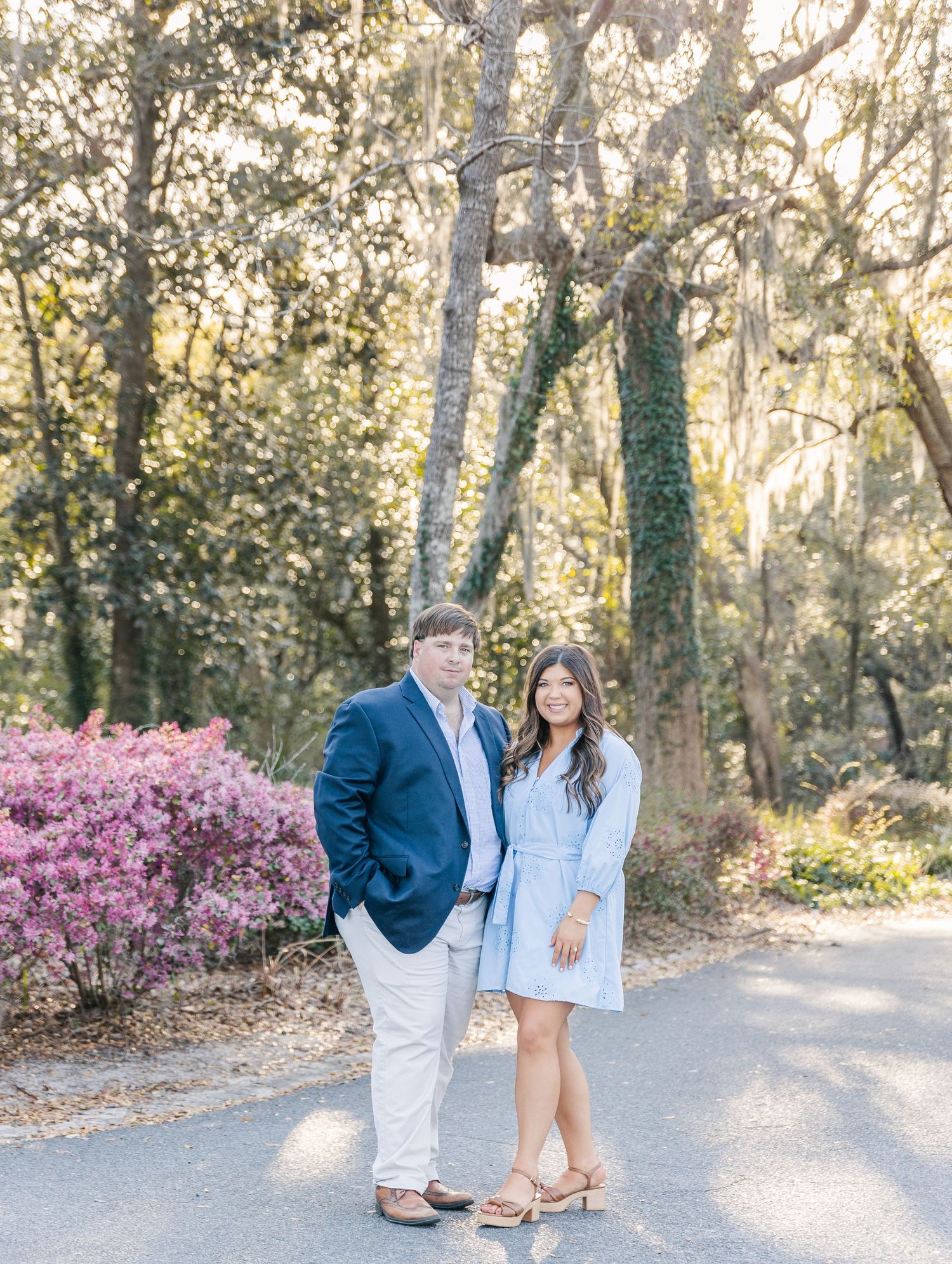 A couple stands together on a path with blooming azaleas and tall trees in the background.