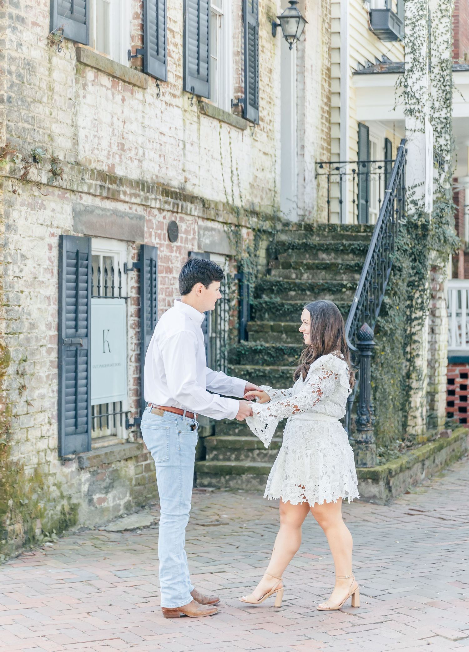A couple holds hands on a brick path in front of historic architecture with blue shutters.