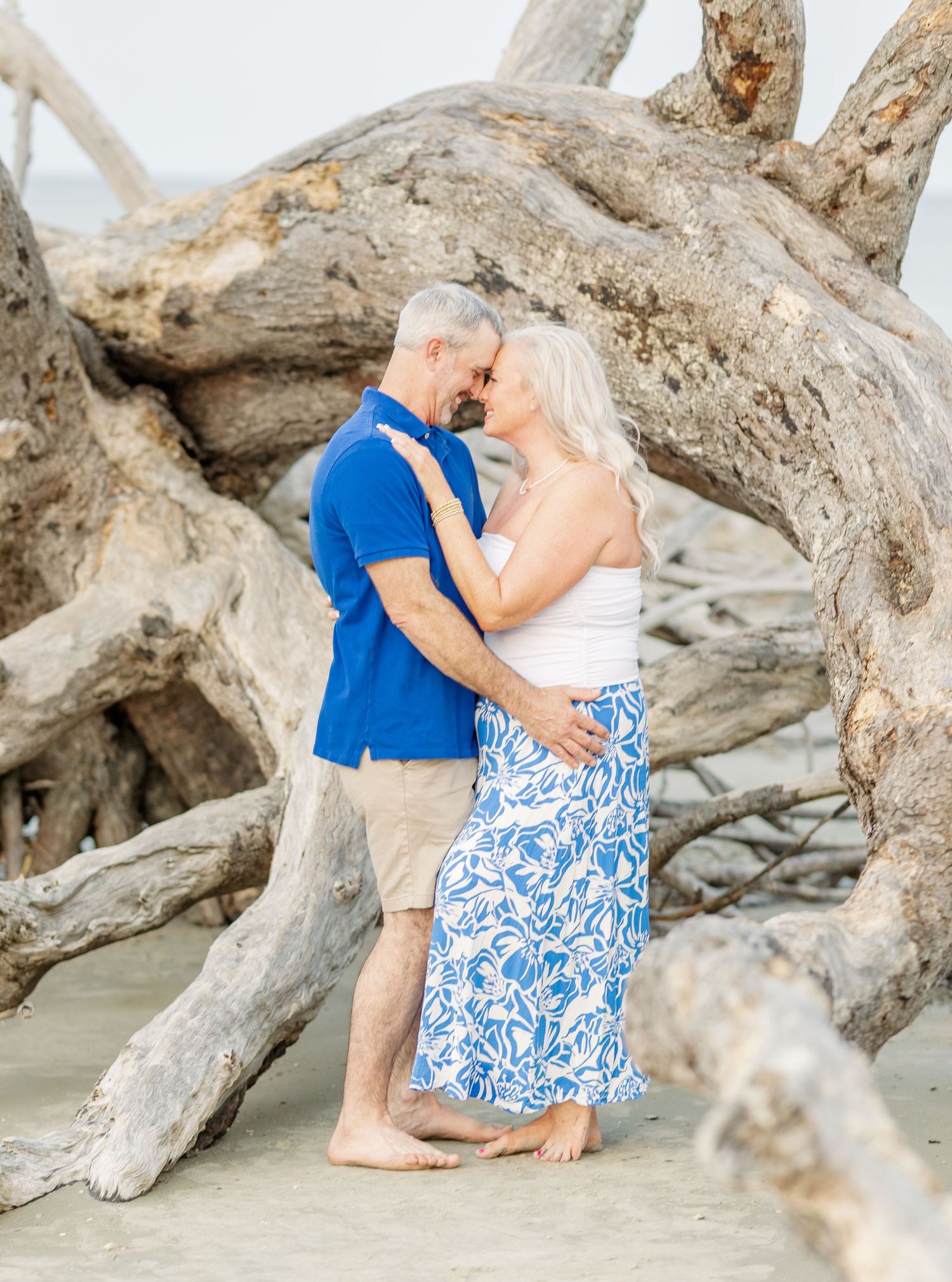 Romantic beach portrait with driftwood backdrop featuring coordinated blue and white outfits on sandy shore.