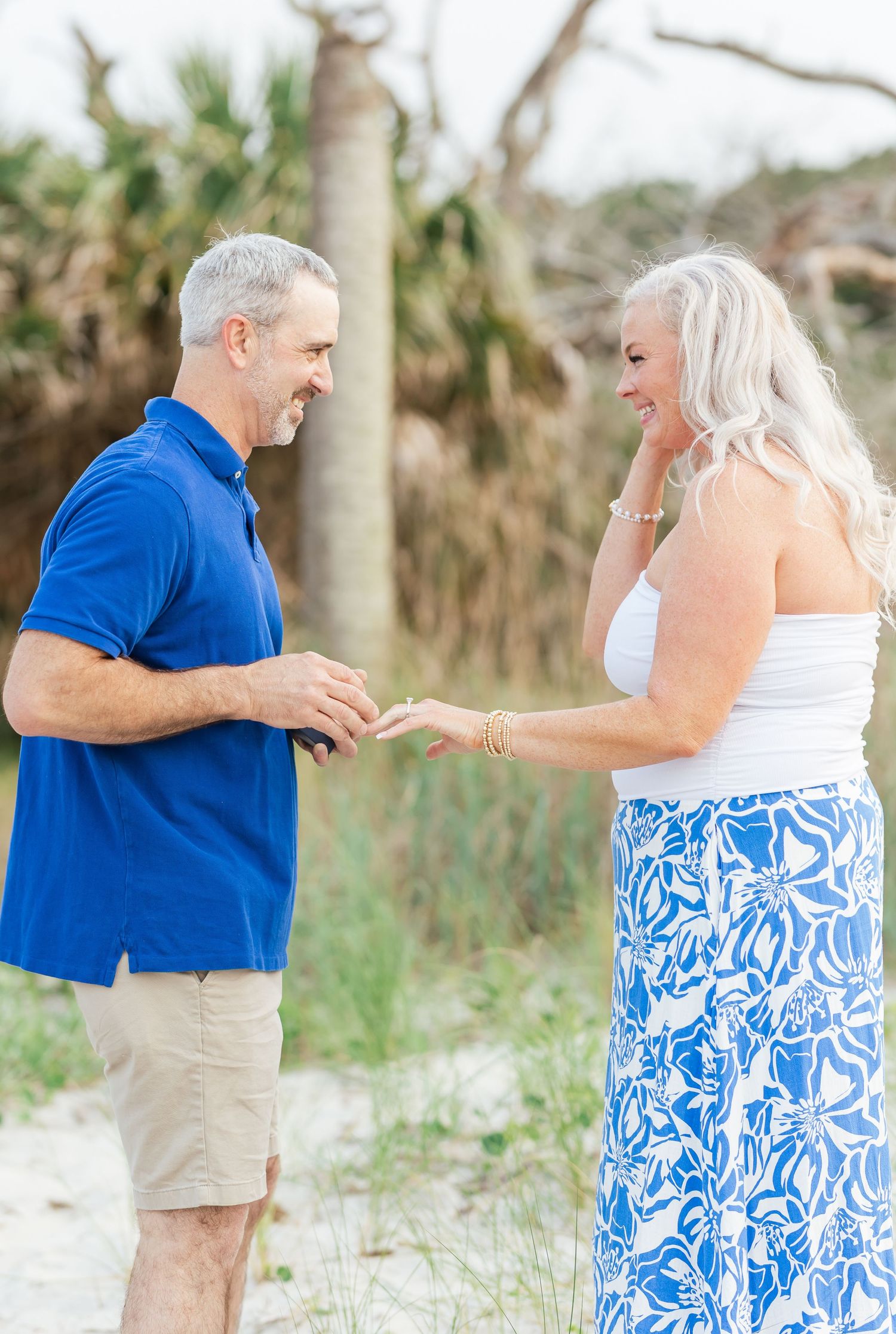 A couple in coordinating blue and white outfits stands together on a sandy beach path surrounded by tropical foliage.