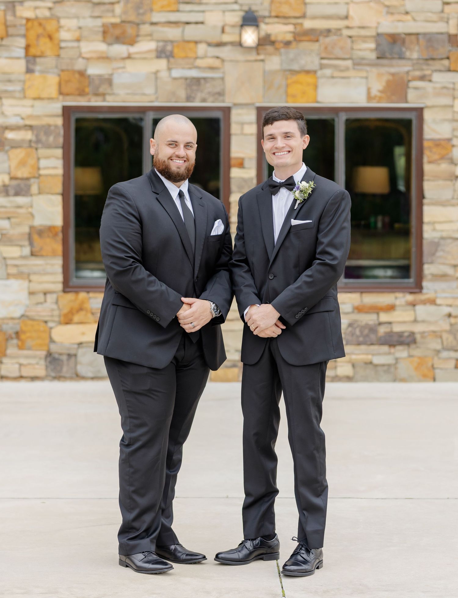 Groomsmen in black tuxedos pose together against a stone wall backdrop at a formal event.