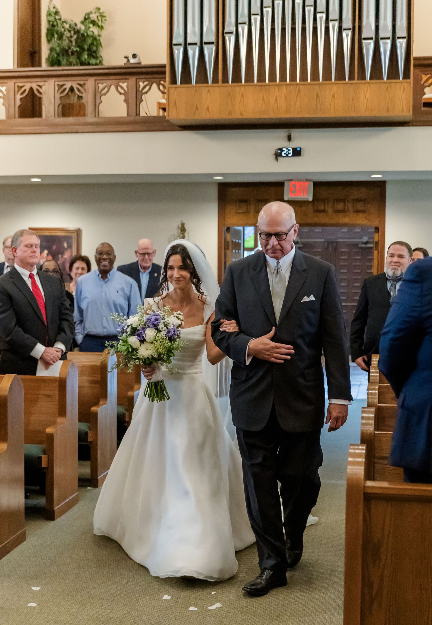 Wedding ceremony processional in traditional church with wooden pews and pipe organ.