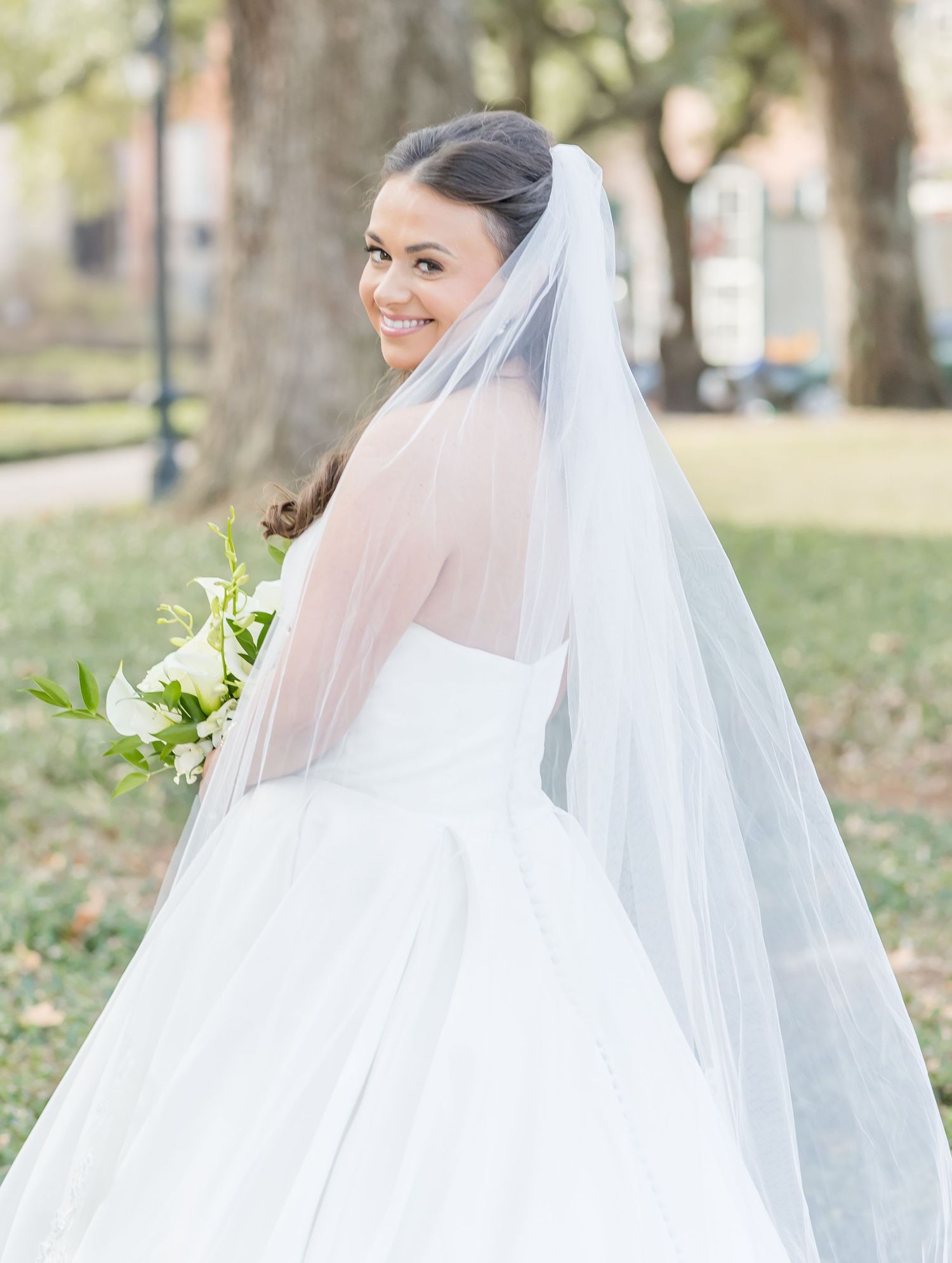 Bride in flowing white veil holds classic white bouquet against outdoor garden backdrop.