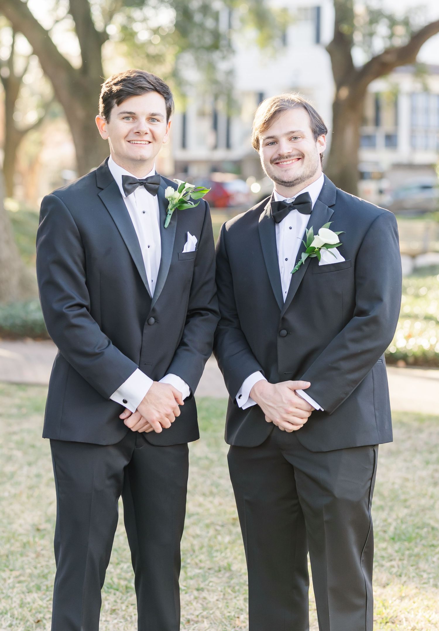 Groomsmen share laughs while posing in formal black tuxedos outdoors.