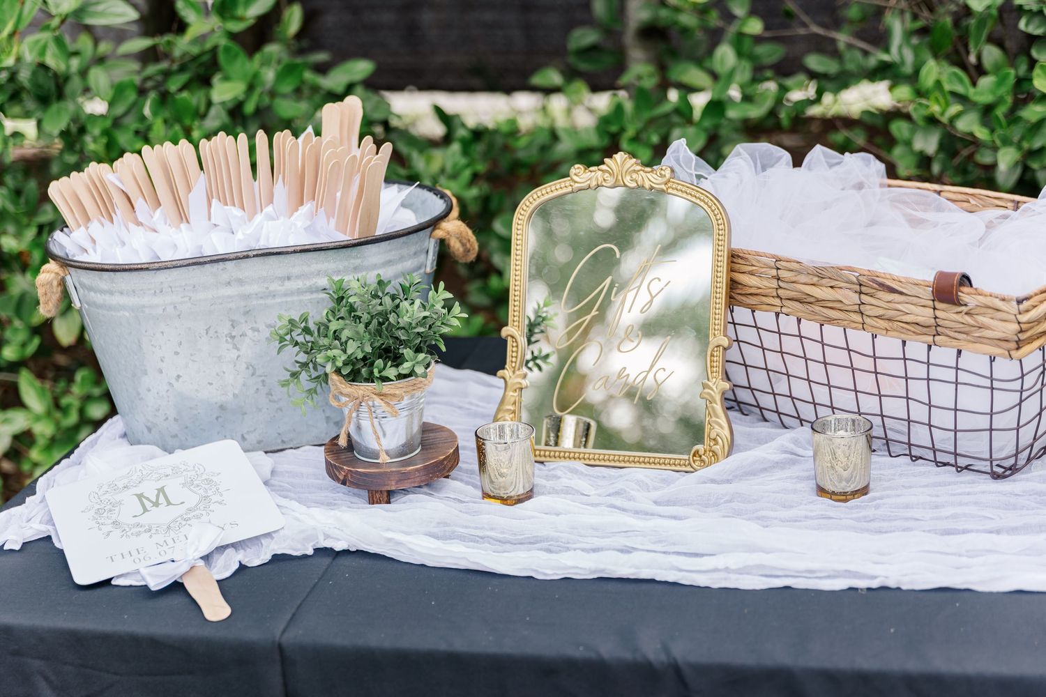 A rustic outdoor wedding table setting with white linens, greenery, and vintage decor elements in a garden setting.