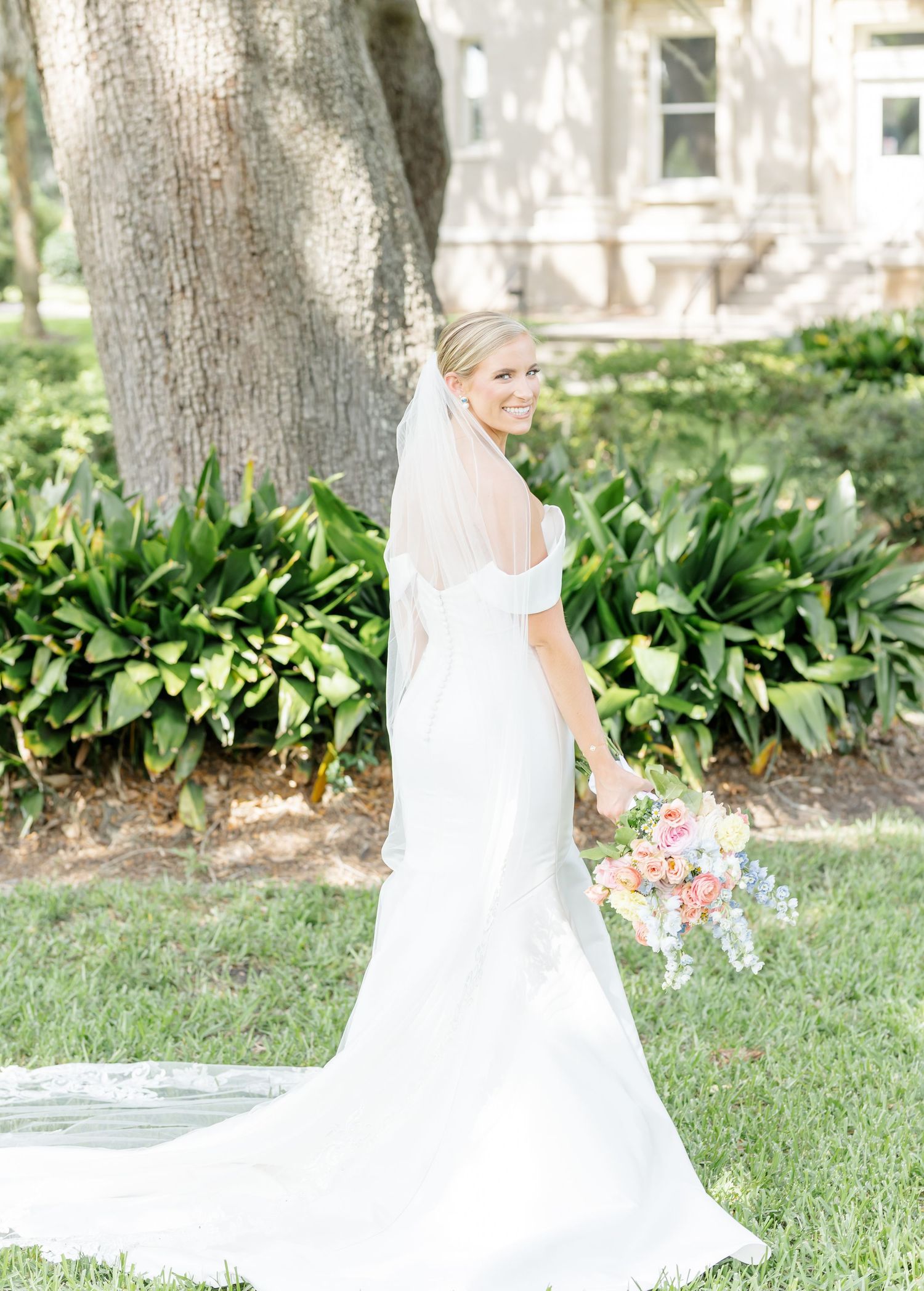 A bride in a flowing white dress poses gracefully in a garden setting with lush greenery and trees in the background.