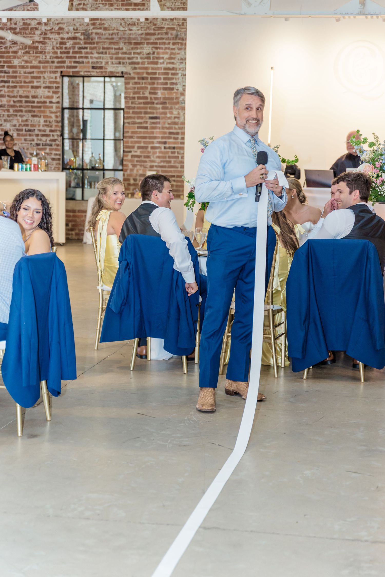 Wedding reception guests in blue attire seated at tables in an industrial brick venue with exposed walls.