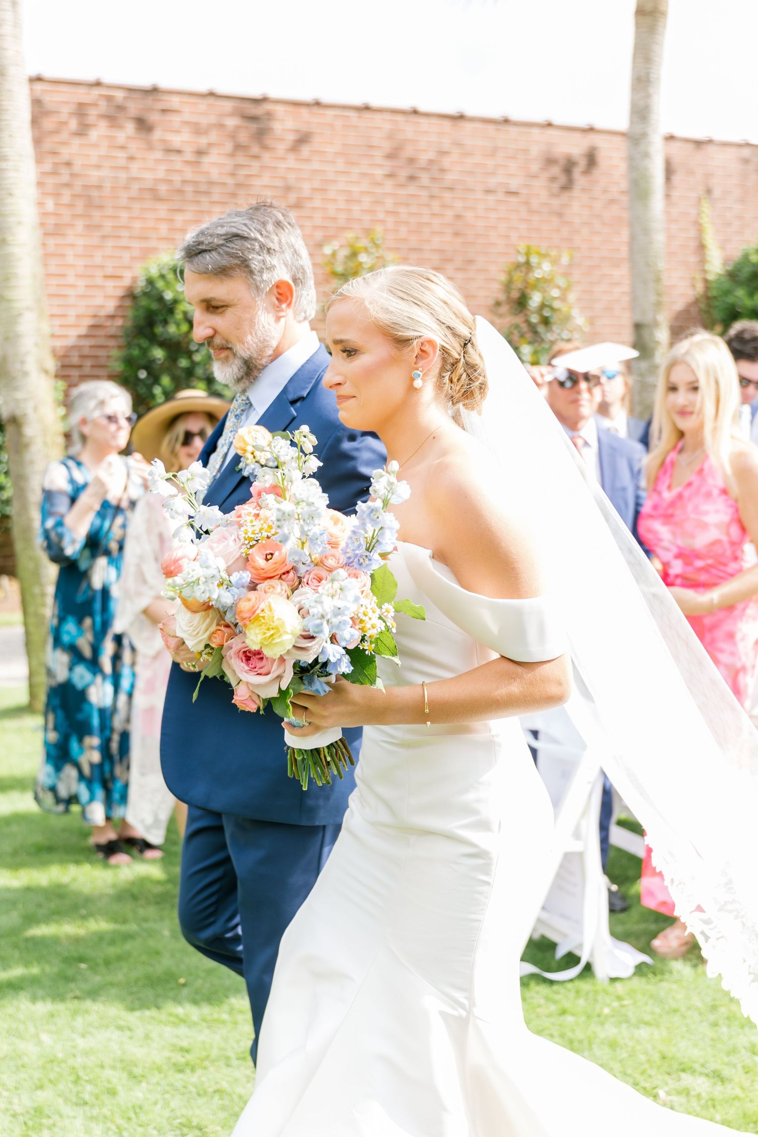 A wedding party celebrates outdoors on a sunny day with pastel floral bouquets near a brick building.