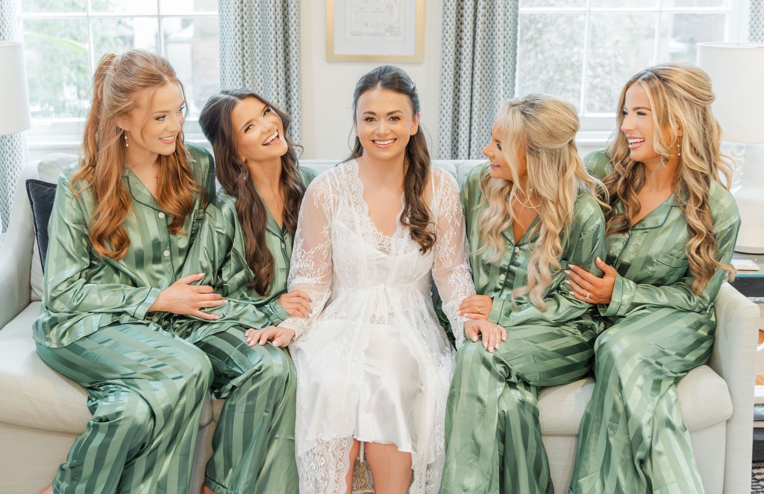 Bridesmaids in sage green silk robes gather around bride in white robe during wedding day preparations.
