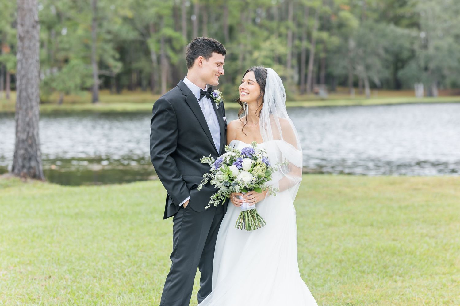 A bride and groom share an intimate moment outdoors near a lake with tall pine trees in the background.