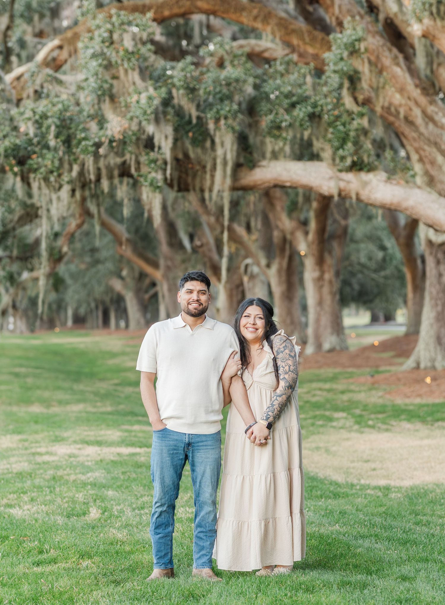 A couple poses together on a grassy lawn beneath towering oak trees draped with Spanish moss.