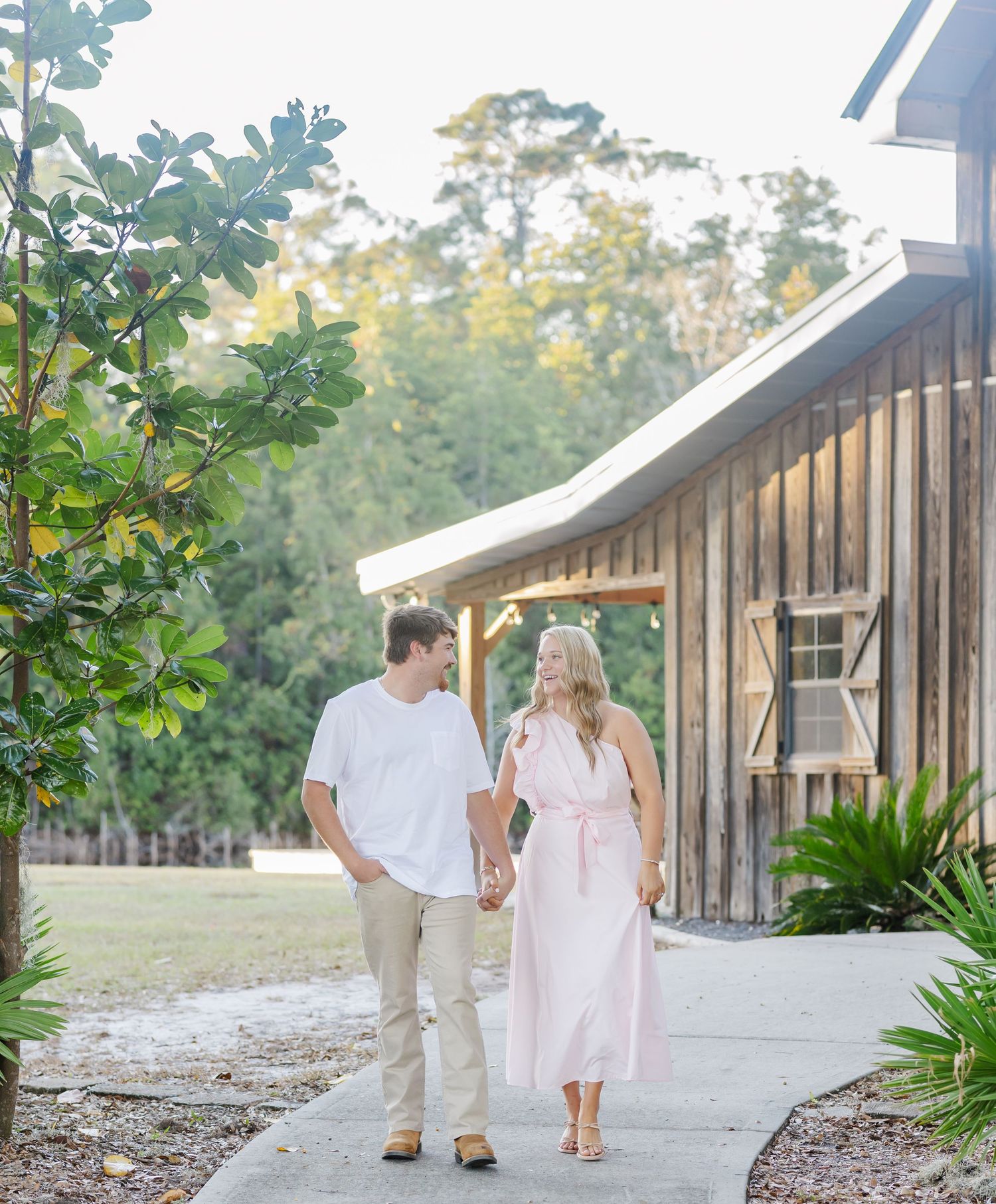 A couple strolls hand in hand along a pathway beside a rustic wooden barn building surrounded by greenery.