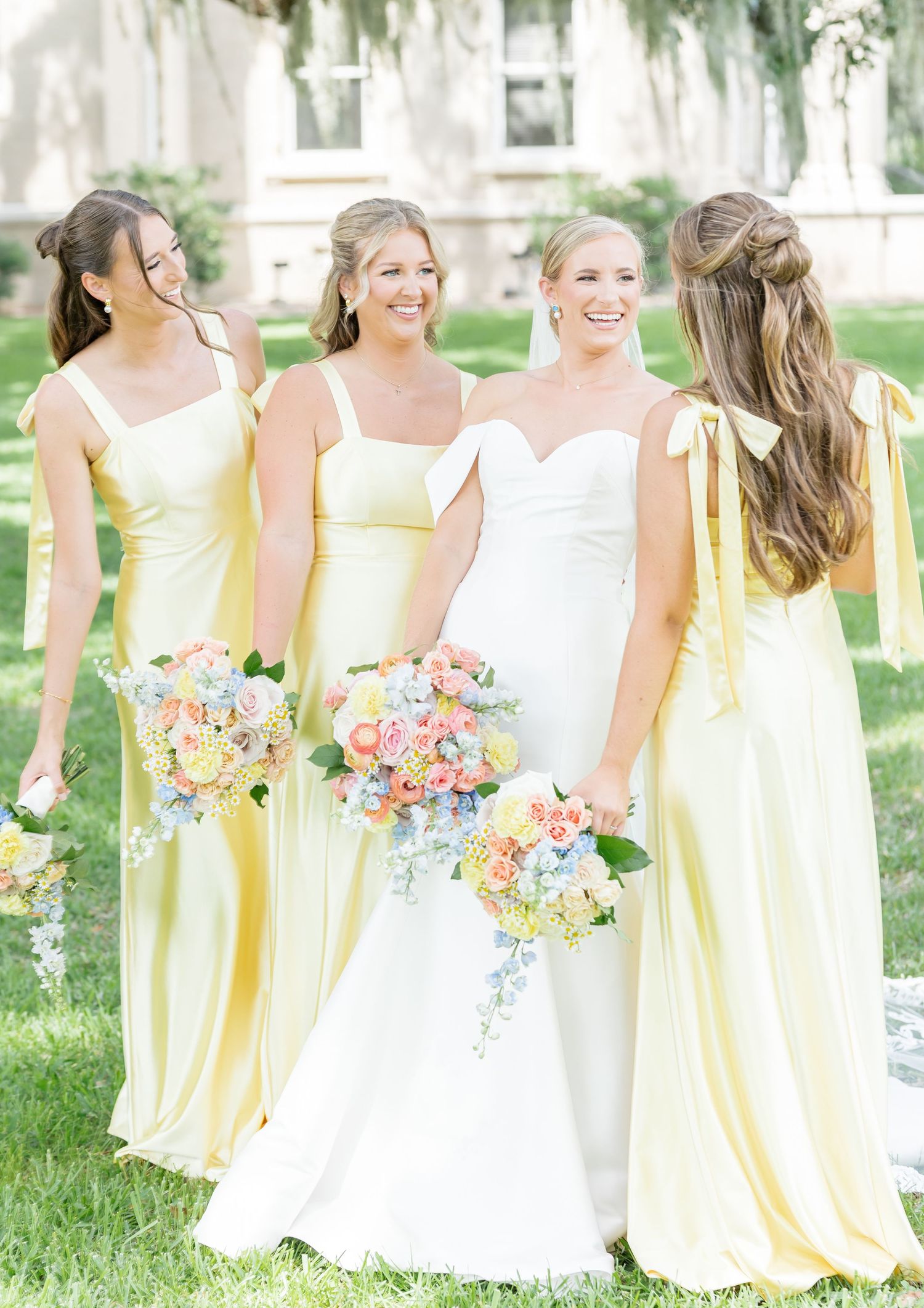 Bridesmaids in pale yellow dresses and bride in white gown pose outdoors with colorful bouquets.