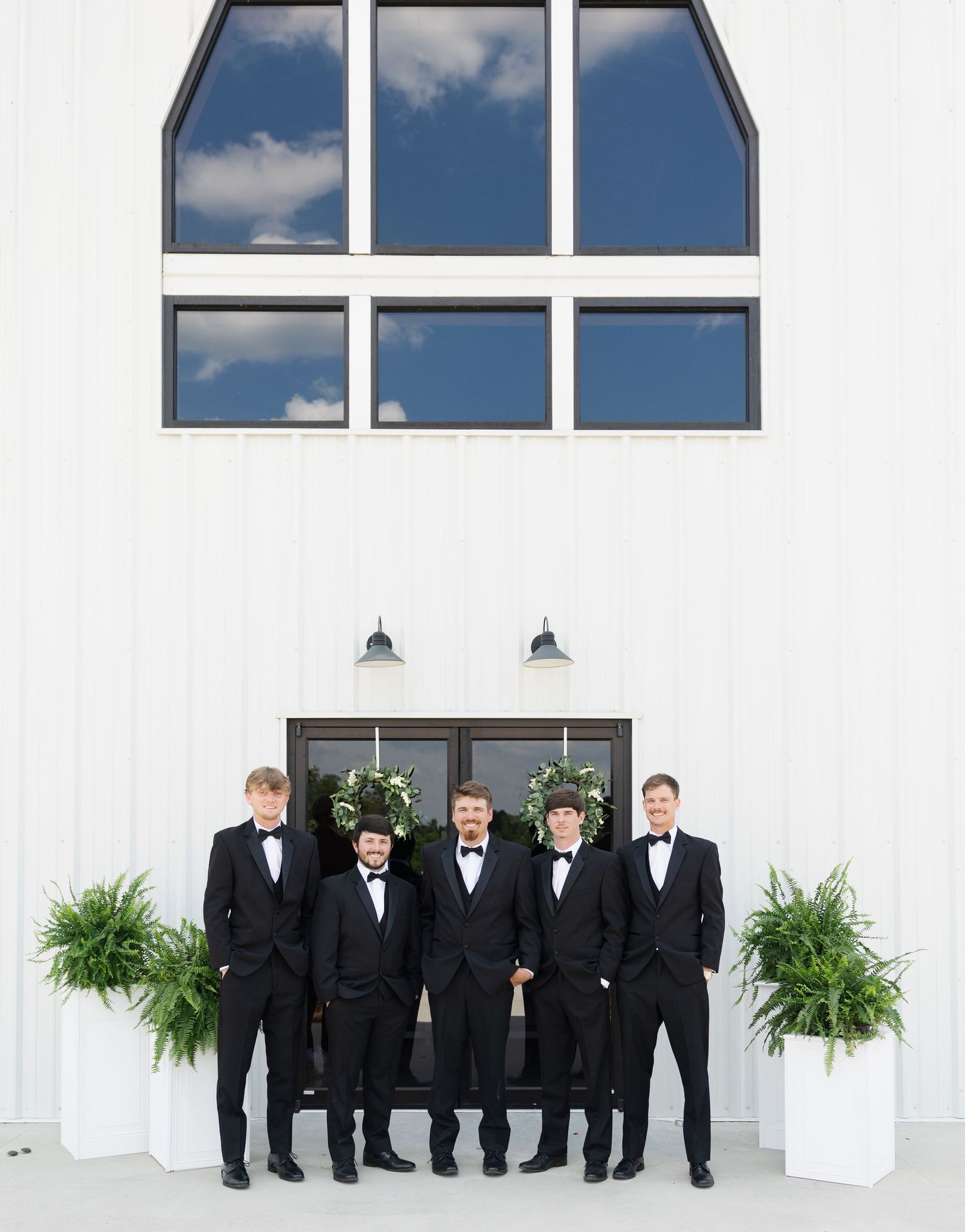 Five groomsmen in black tuxedos pose in front of a white barn venue with greenery wreaths and potted ferns.