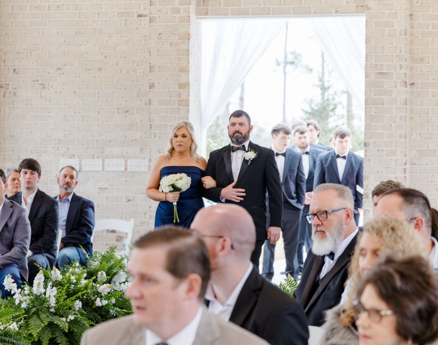 Groomsmen and bridesmaid in navy dress escort guests down aisle in bright white brick ceremony venue.