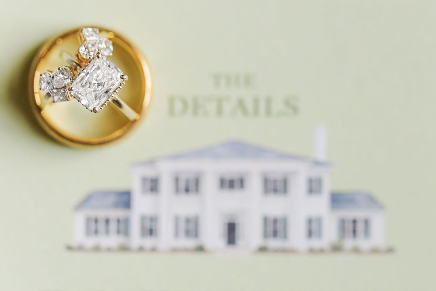 Close-up of a diamond engagement ring in a gold dish atop elegant wedding stationery with a venue illustration.