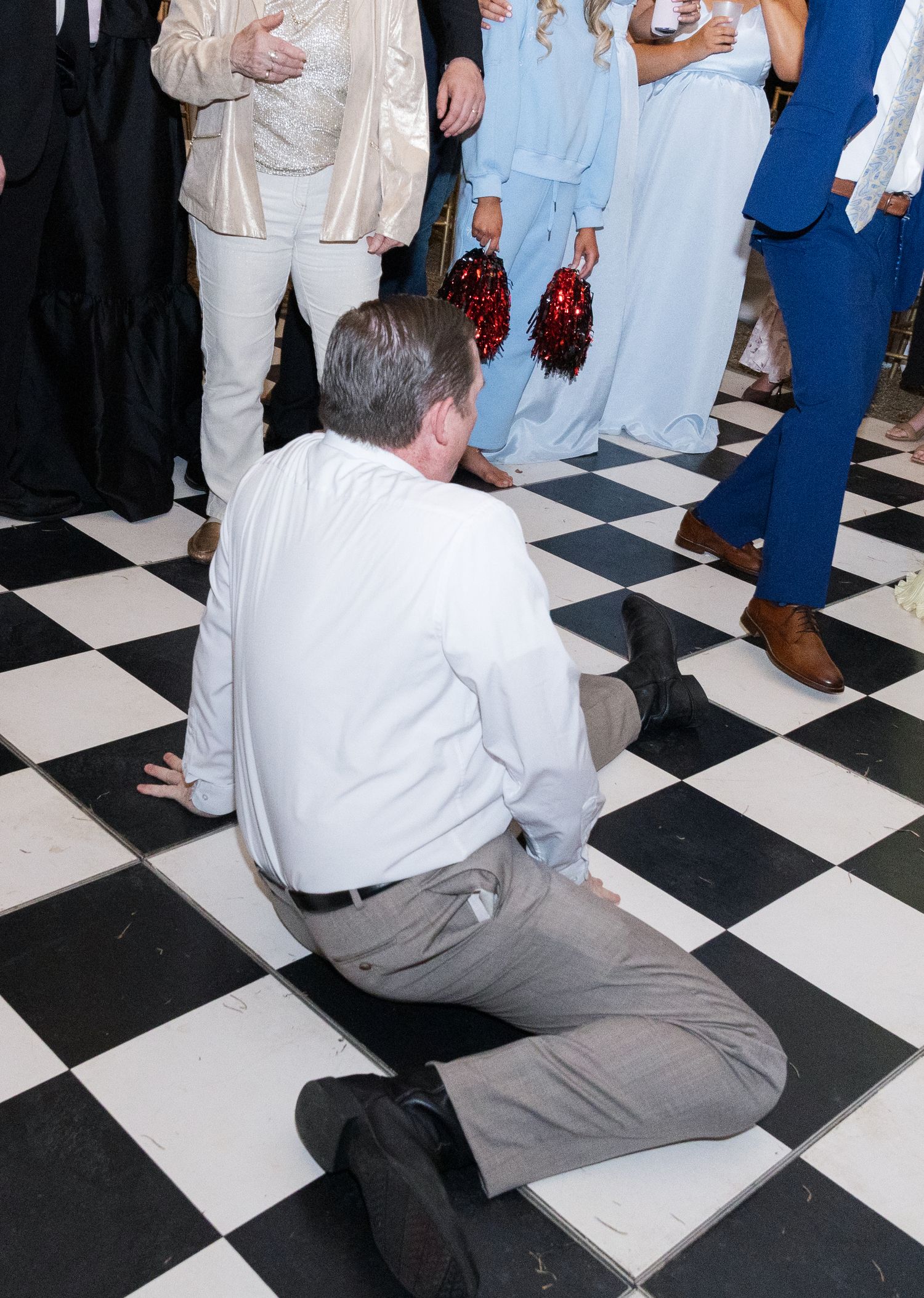 Man kneeling on black and white checkered dance floor surrounded by wedding guests during reception.